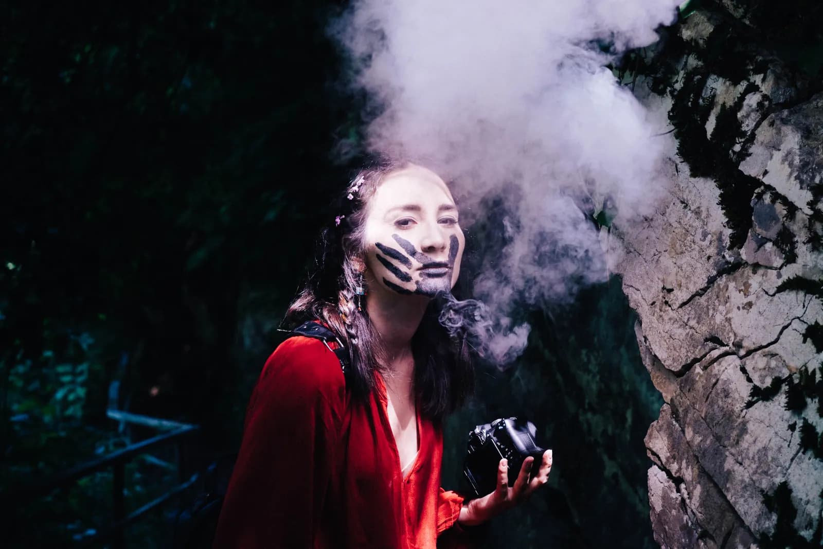 Woman in a red dress with a painted handprint on her face holding a camera, smoke rising in the dark