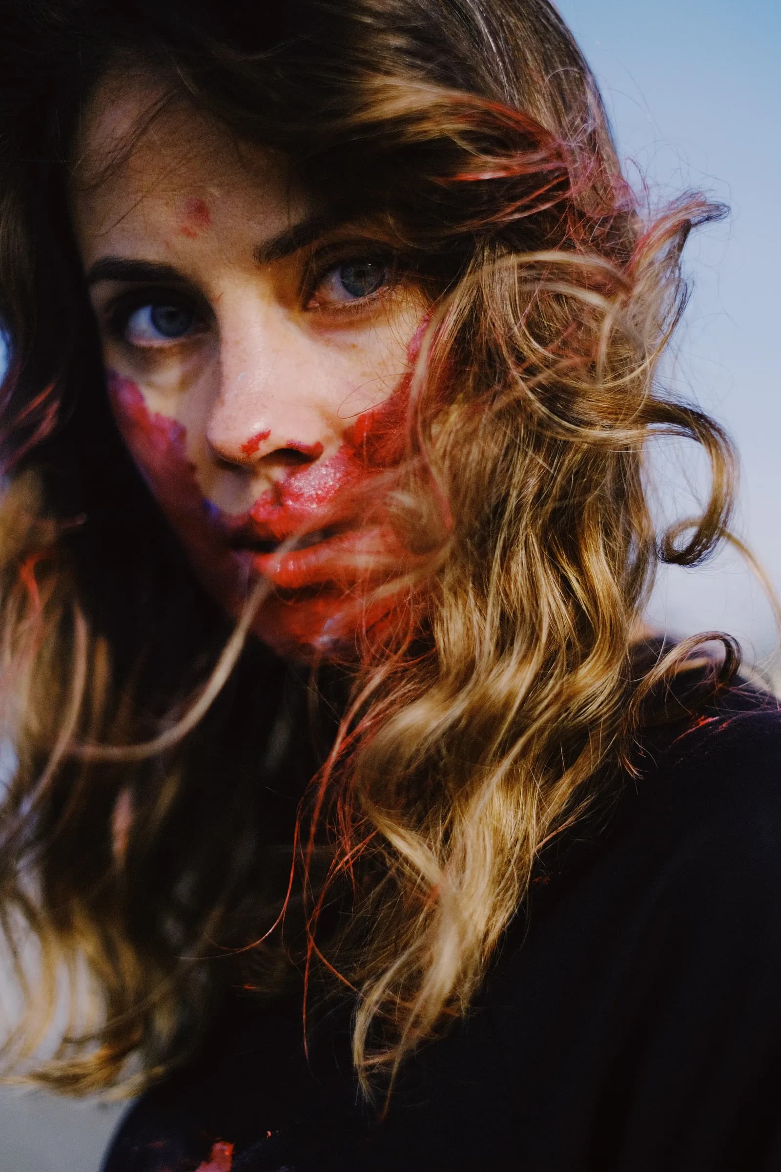 Portrait of a woman with crimson paint across her face, hair blowing against a blue sky