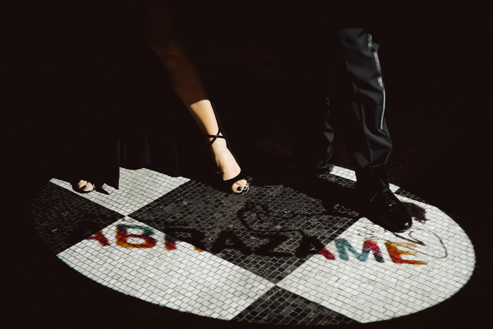 Feet of two tango dancers on a mosaic floor, the word ABRÁZAME projected in a circle of light