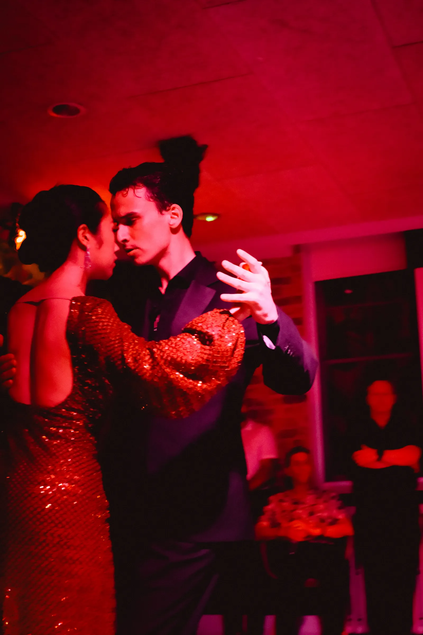 Young couple dancing tango in deep red light, her sequinned dress catching the glow