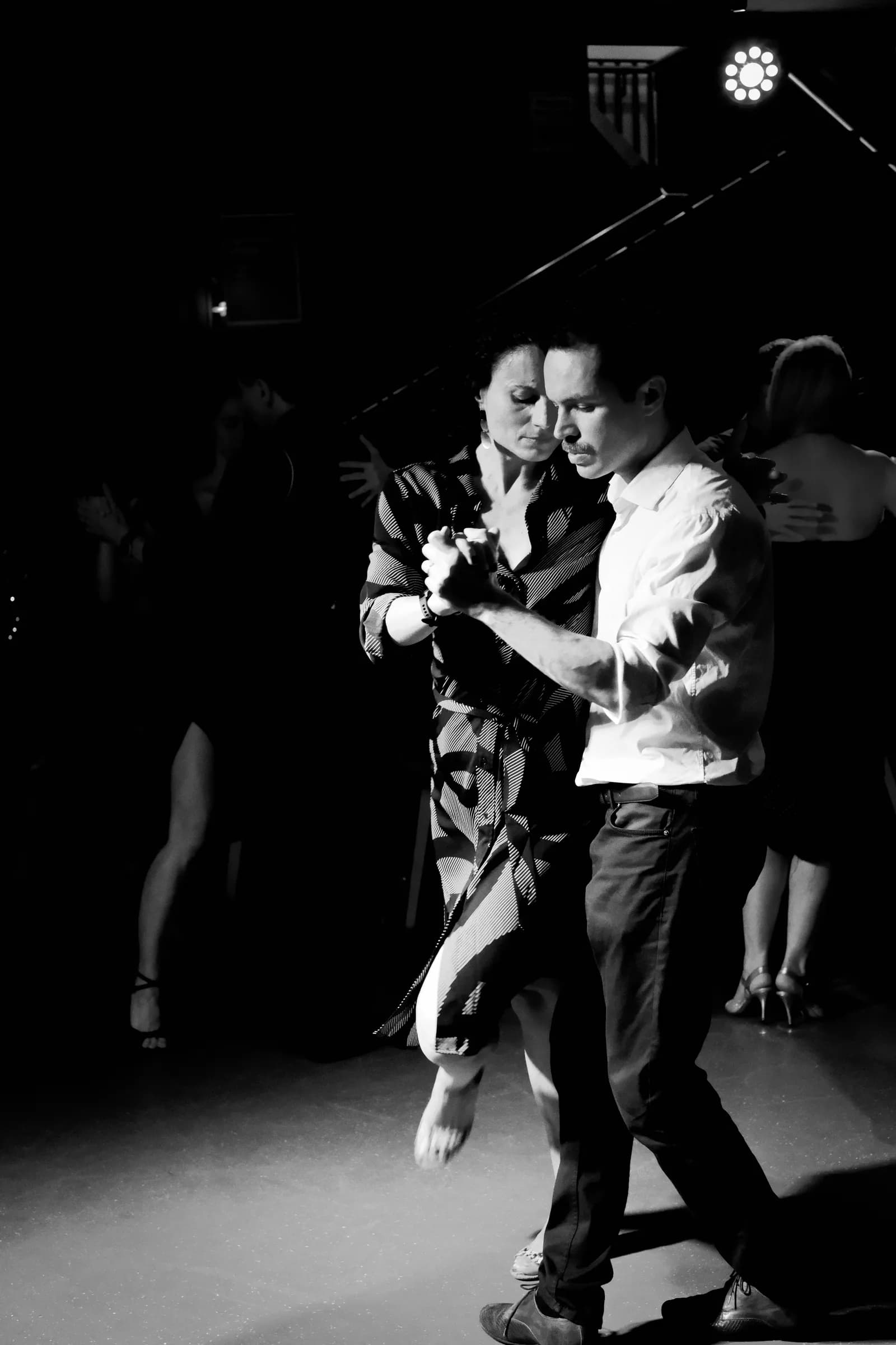 Couple in close embrace dancing at a London milonga, other dancers in the dark behind them