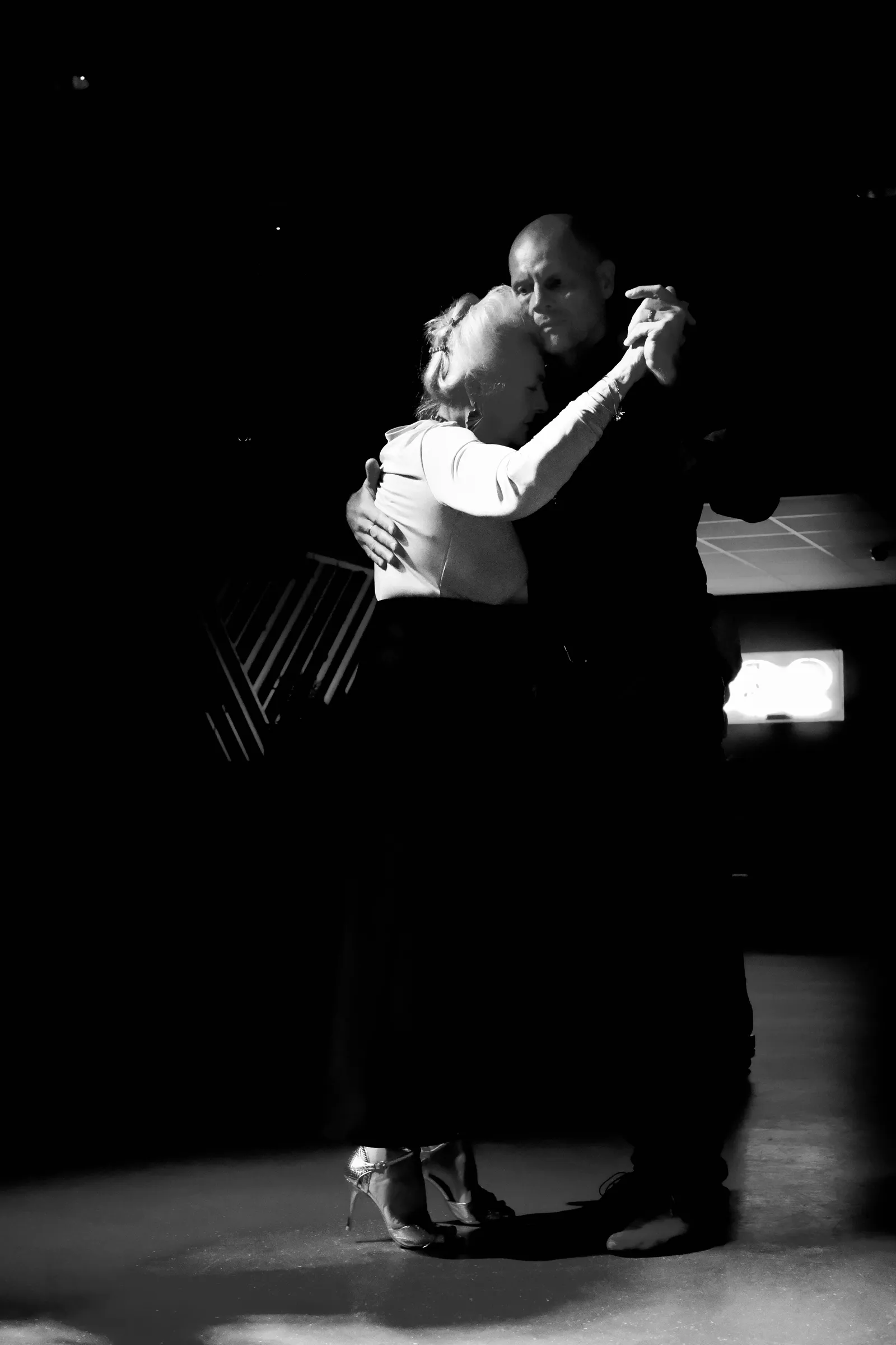Grey-haired couple in a close embrace, lit by a single beam in a dark hall