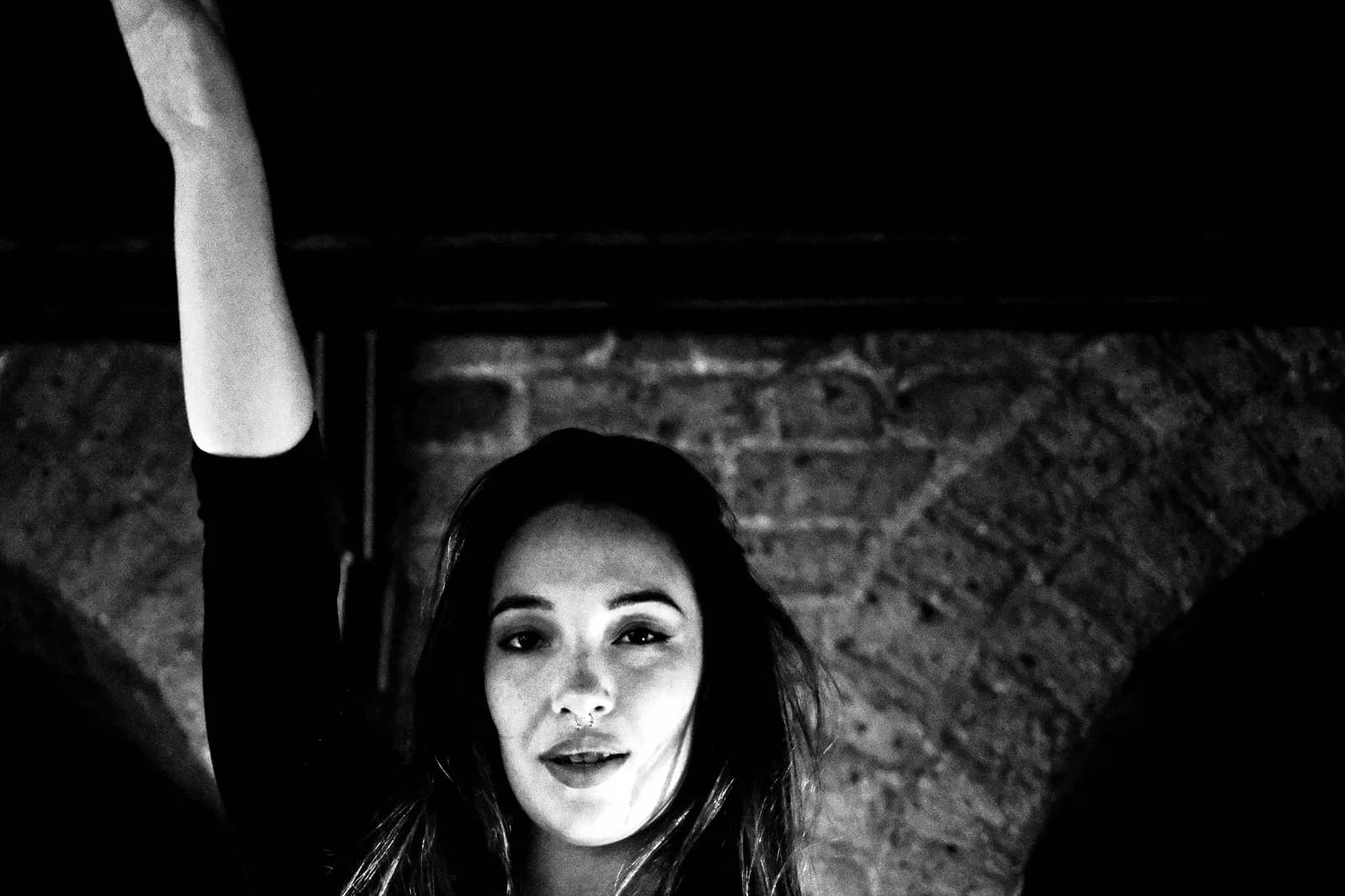Woman looking into the lens mid-dance, one arm raised, brick arches behind her