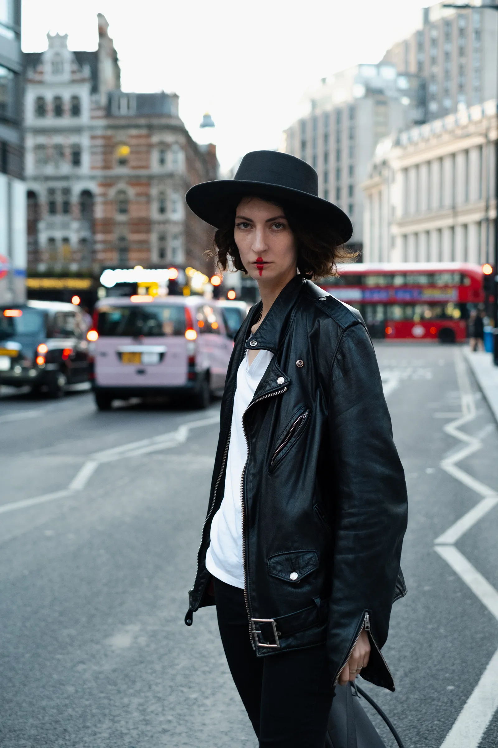 Musician in a leather jacket and black hat standing in a London street, a red double-decker bus behind her