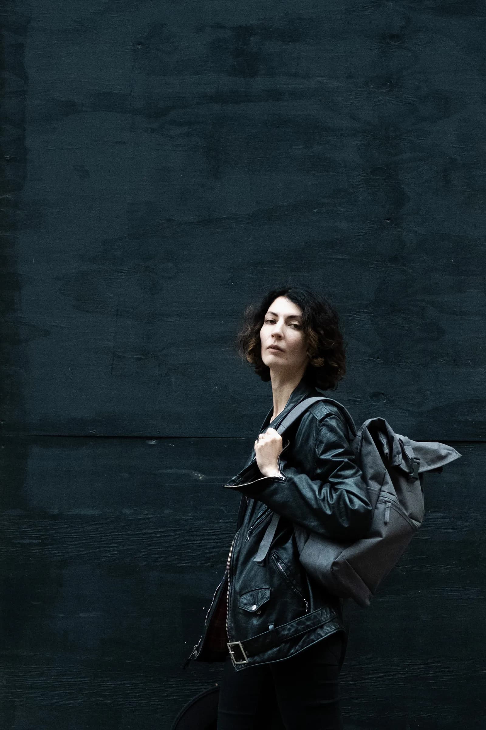 Woman with a leather rucksack glancing back against a dark blue wall