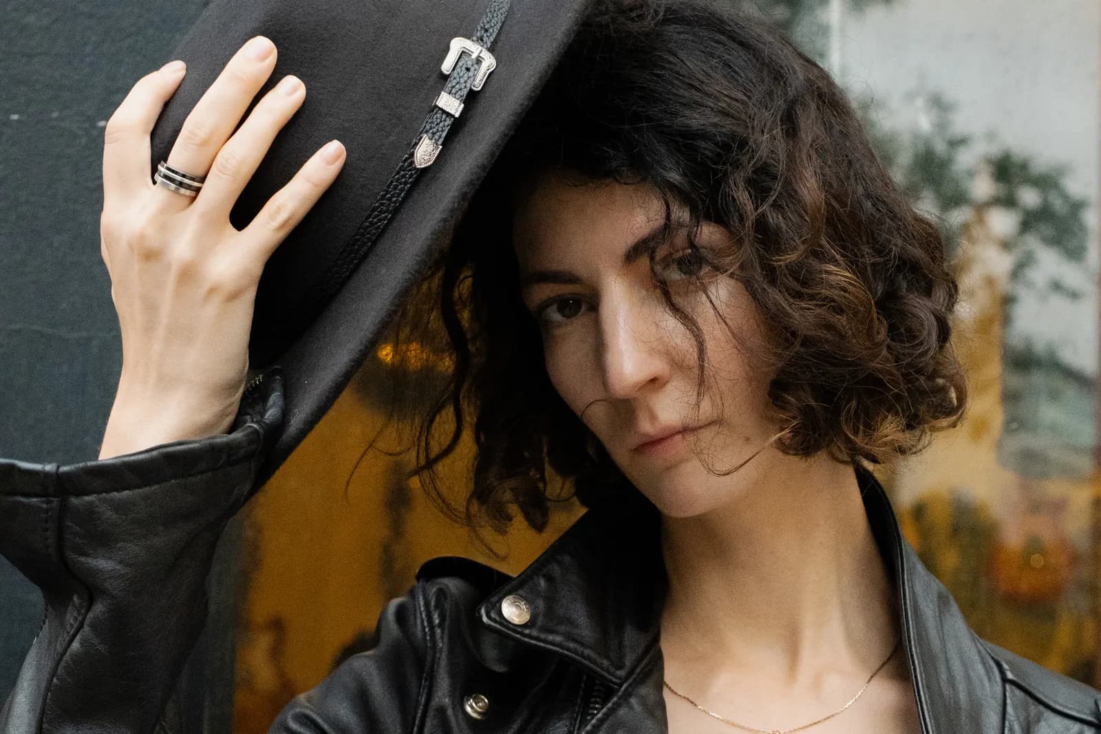 Woman lifting her black hat by the crown, ringed fingers, curls loose