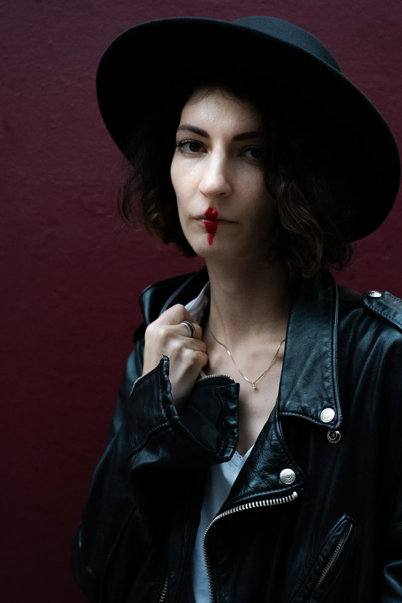 Musician in a hat against a dark red wall, lipstick run down from her lip