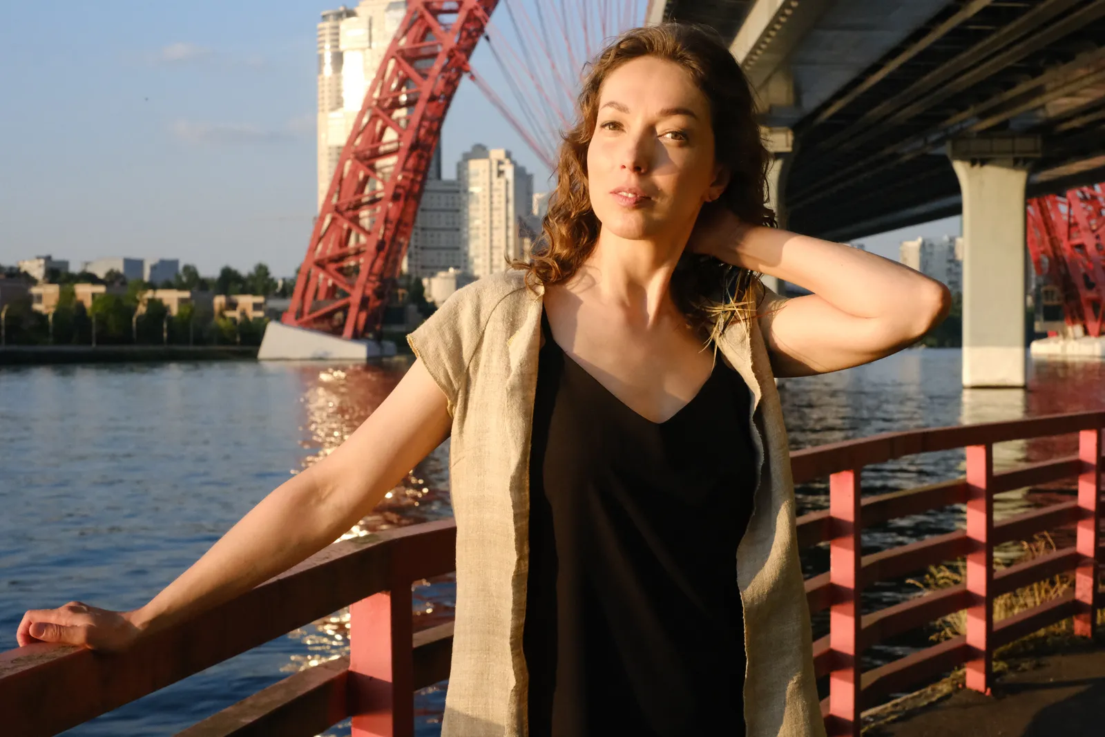 Woman at a red railing under a great red bridge, hand in her hair, evening sun on the water