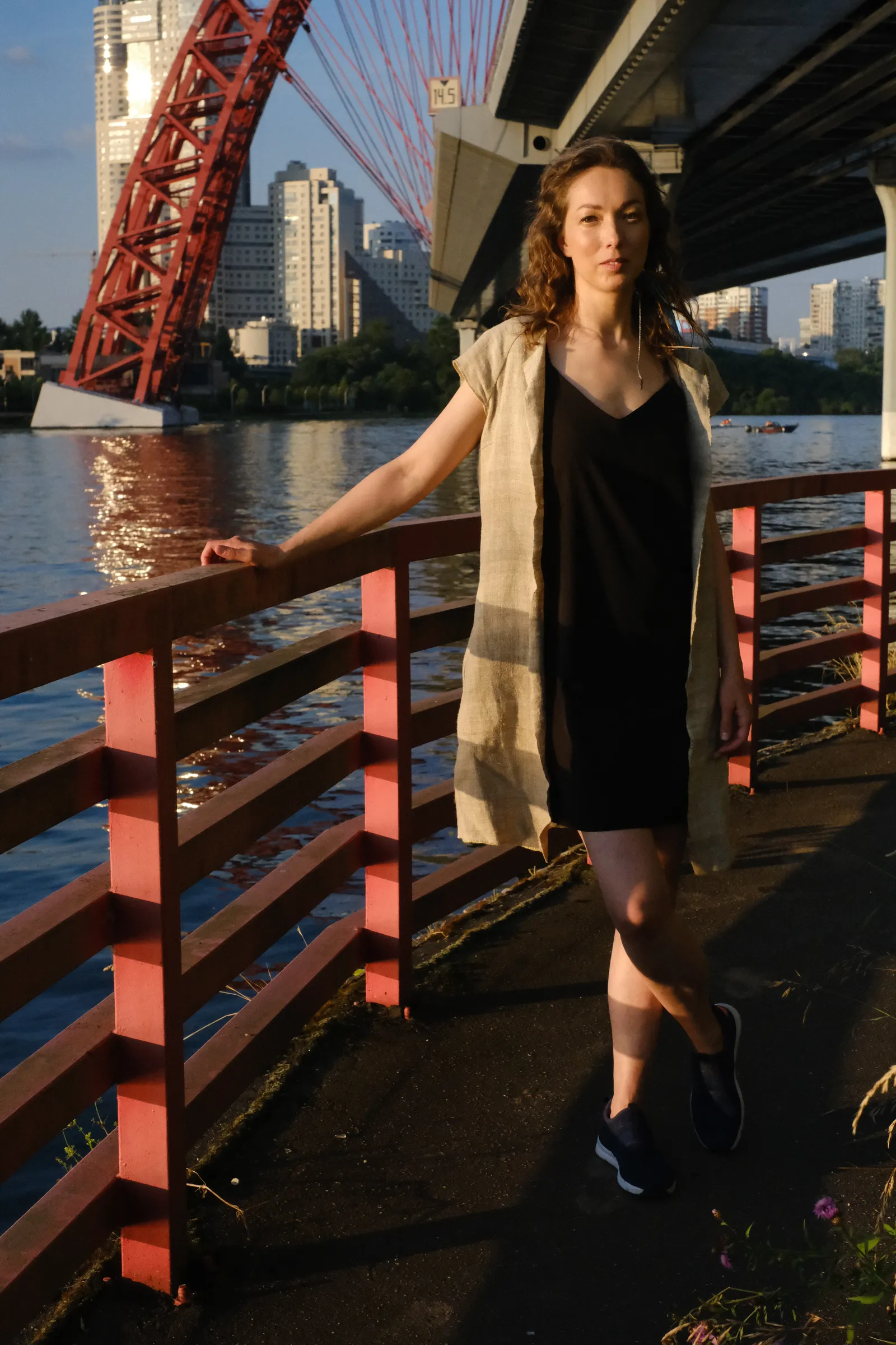 Woman in a black slip dress and linen coat at a riverside railing beneath the bridge