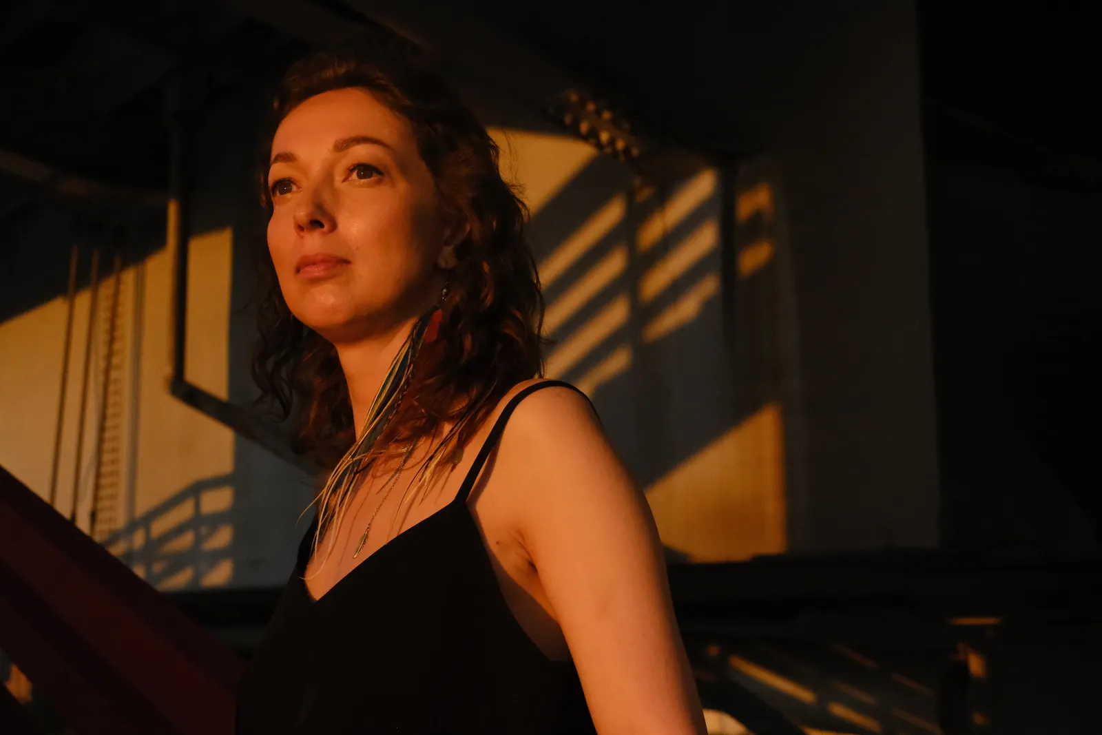 Woman in gold evening light under the bridge girders, cable shadows raked across the wall
