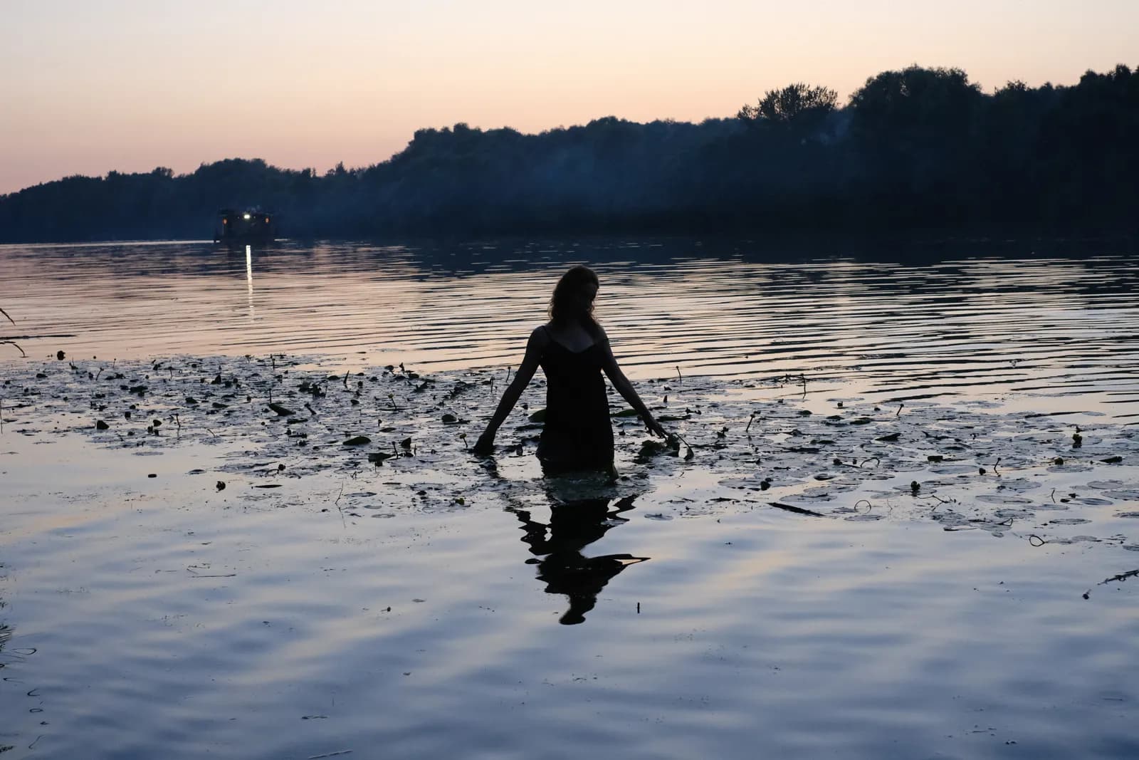 Silhouette of a woman wading into a still river at sunset among the weeds