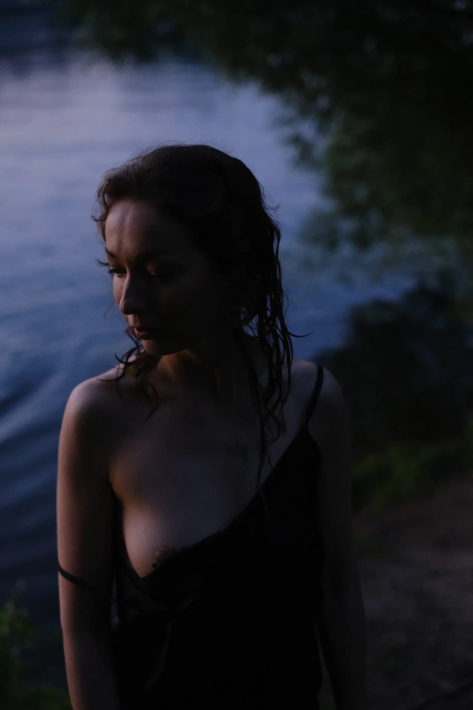 Woman with soaked hair at the water's edge, eyes lowered, the river dark behind
