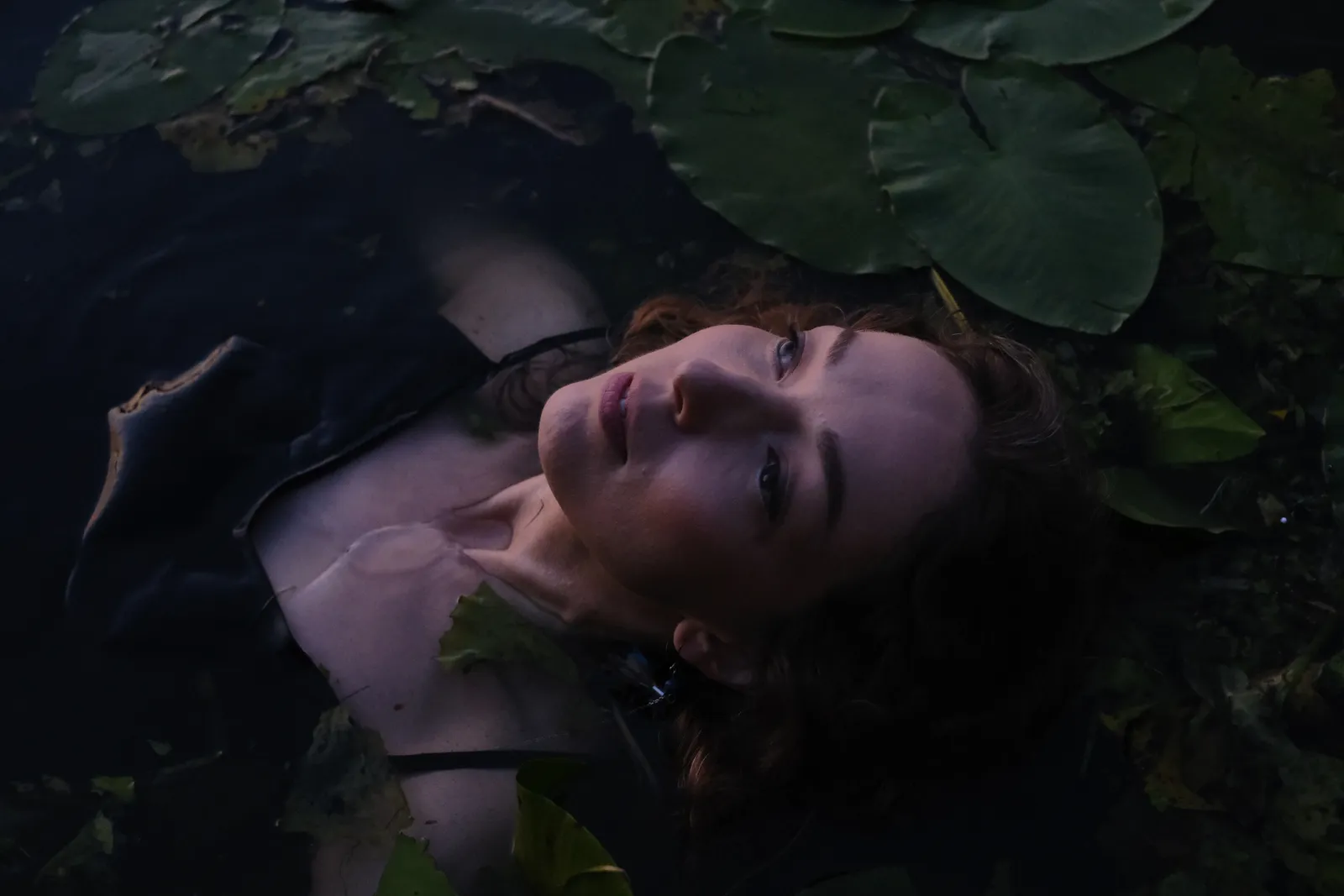 Woman lying in dark water among lily pads, face turned to the sky