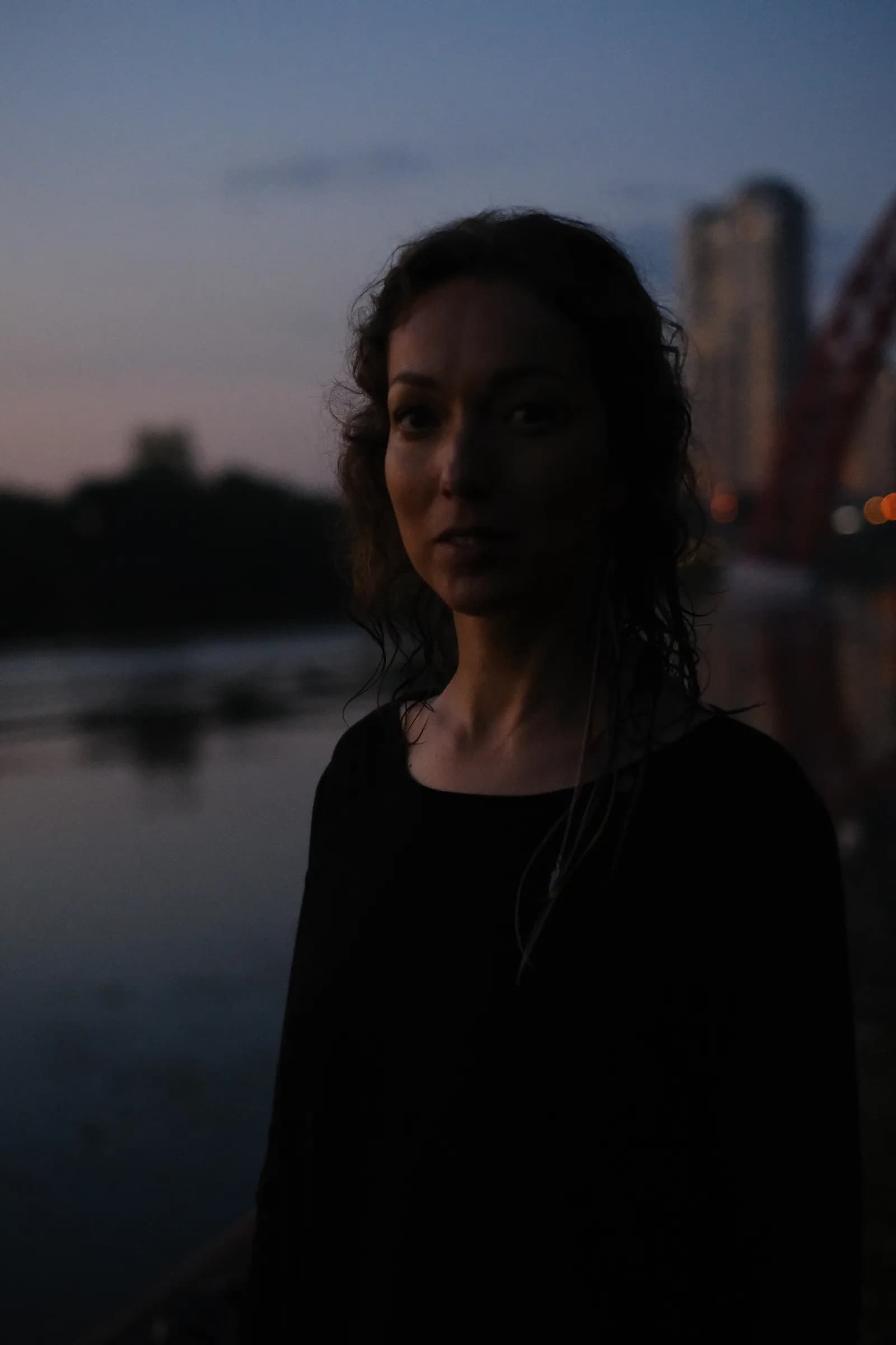 Woman with damp hair facing the lens in deep dusk, river and bridge lights behind