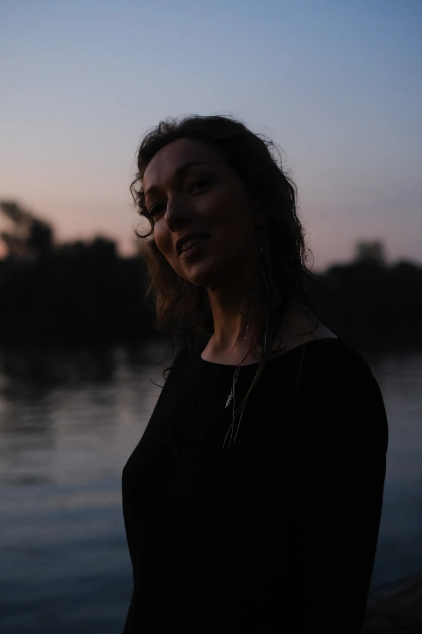 Woman tilting her head at the night river, the sky still pink at the treeline