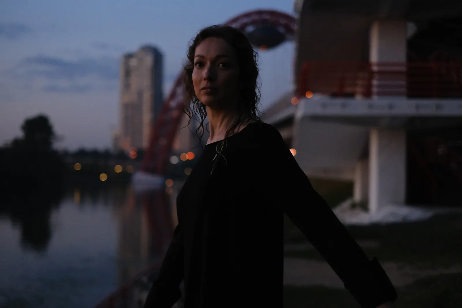 Woman in black at the dusk river, tower lights on across the water