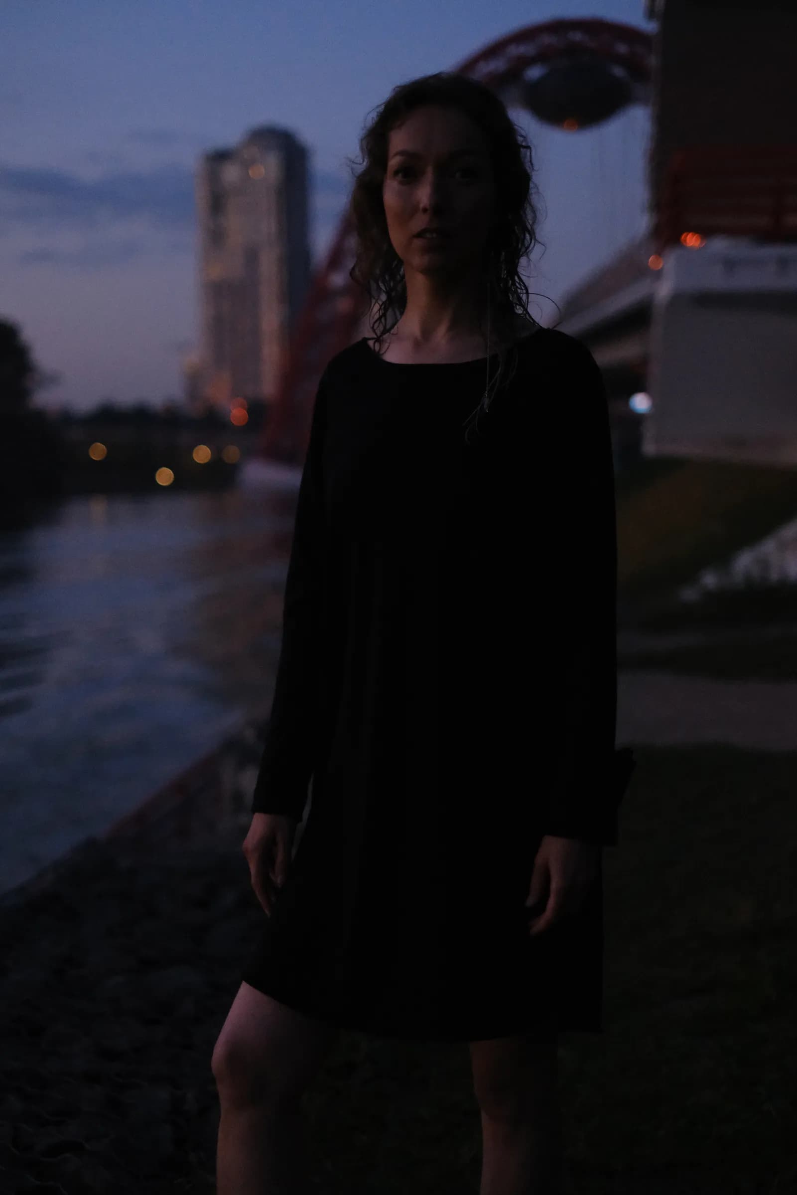 Woman in a loose black dress on the darkening embankment, towers lit across the river