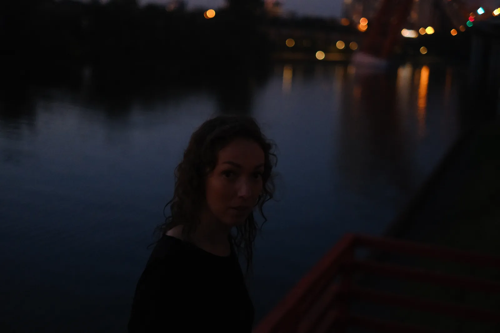 Woman at the red rail above the black river, bridge lights scattered on the water