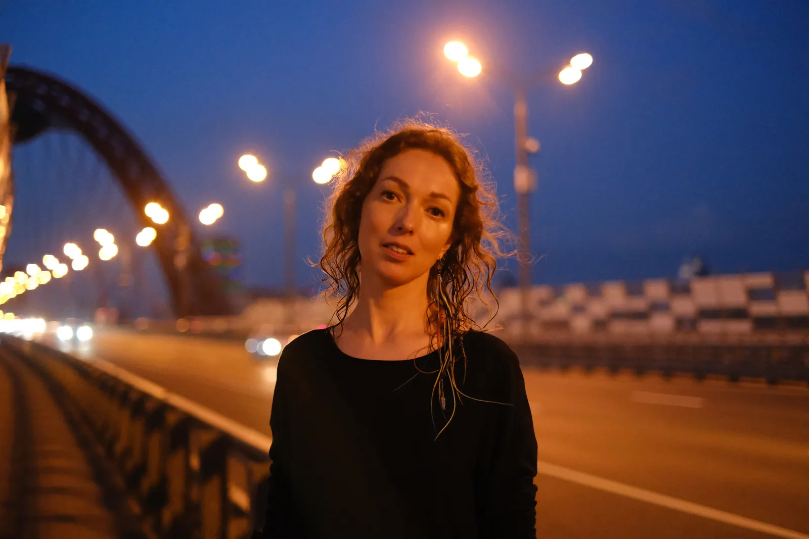 Woman on the bridge roadway at night, the lit arch curving away behind her
