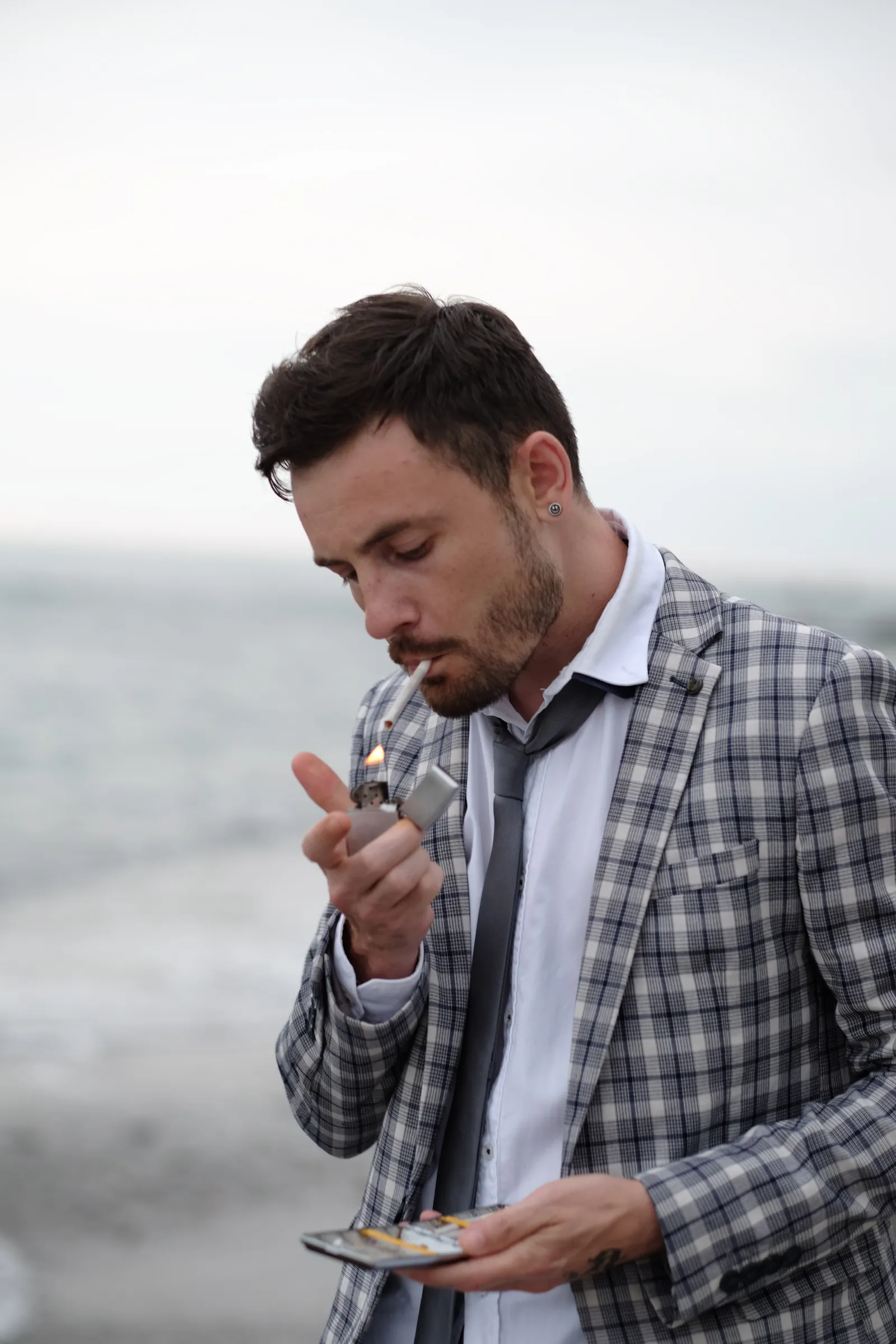 Man in a checked jacket lighting a cigarette against a pale sea