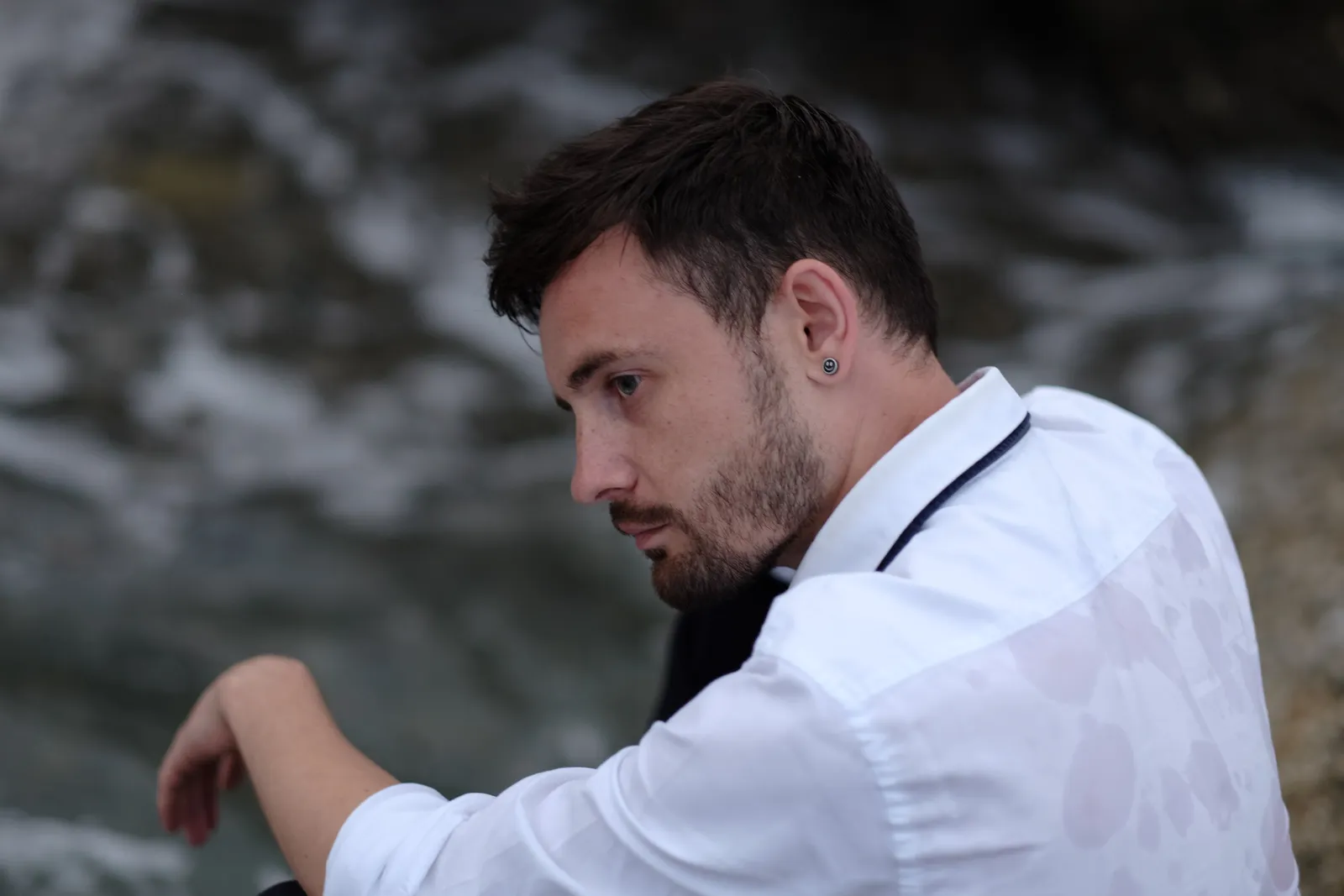 Man in a white shirt looking down over rocks, breaking water blurred behind