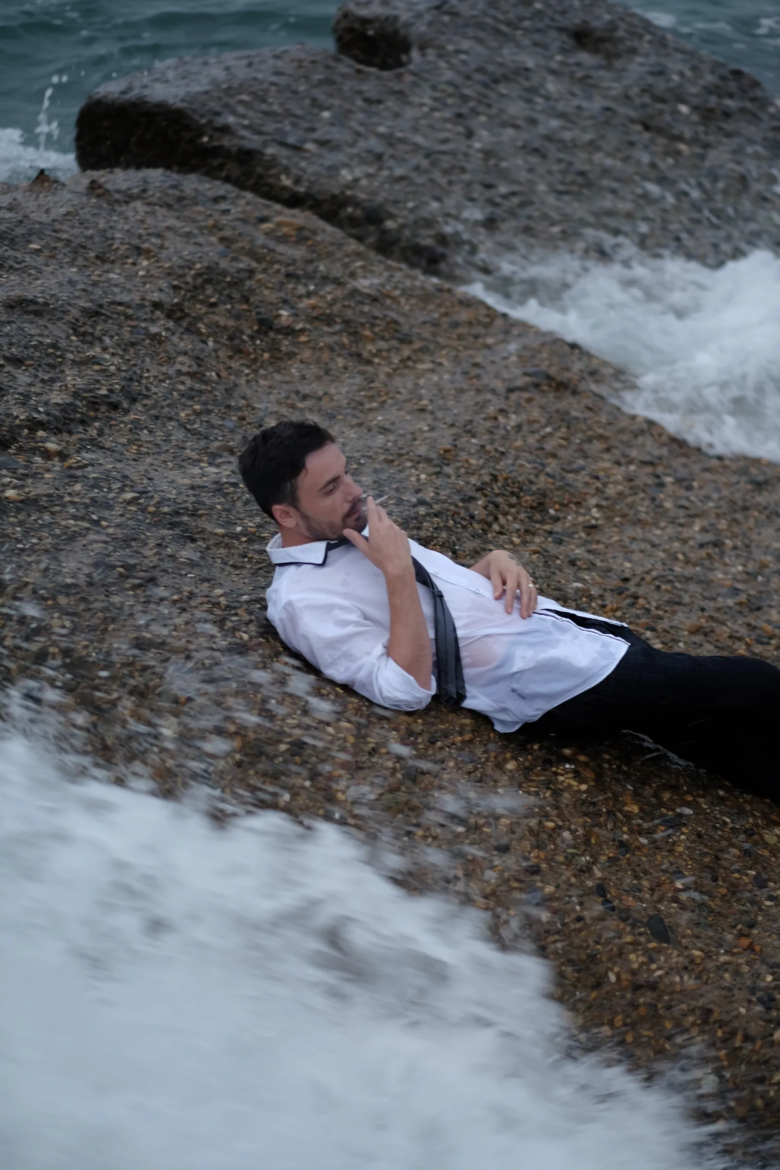 Man in a loose tie lying back on a wet rock as a wave rushes past below