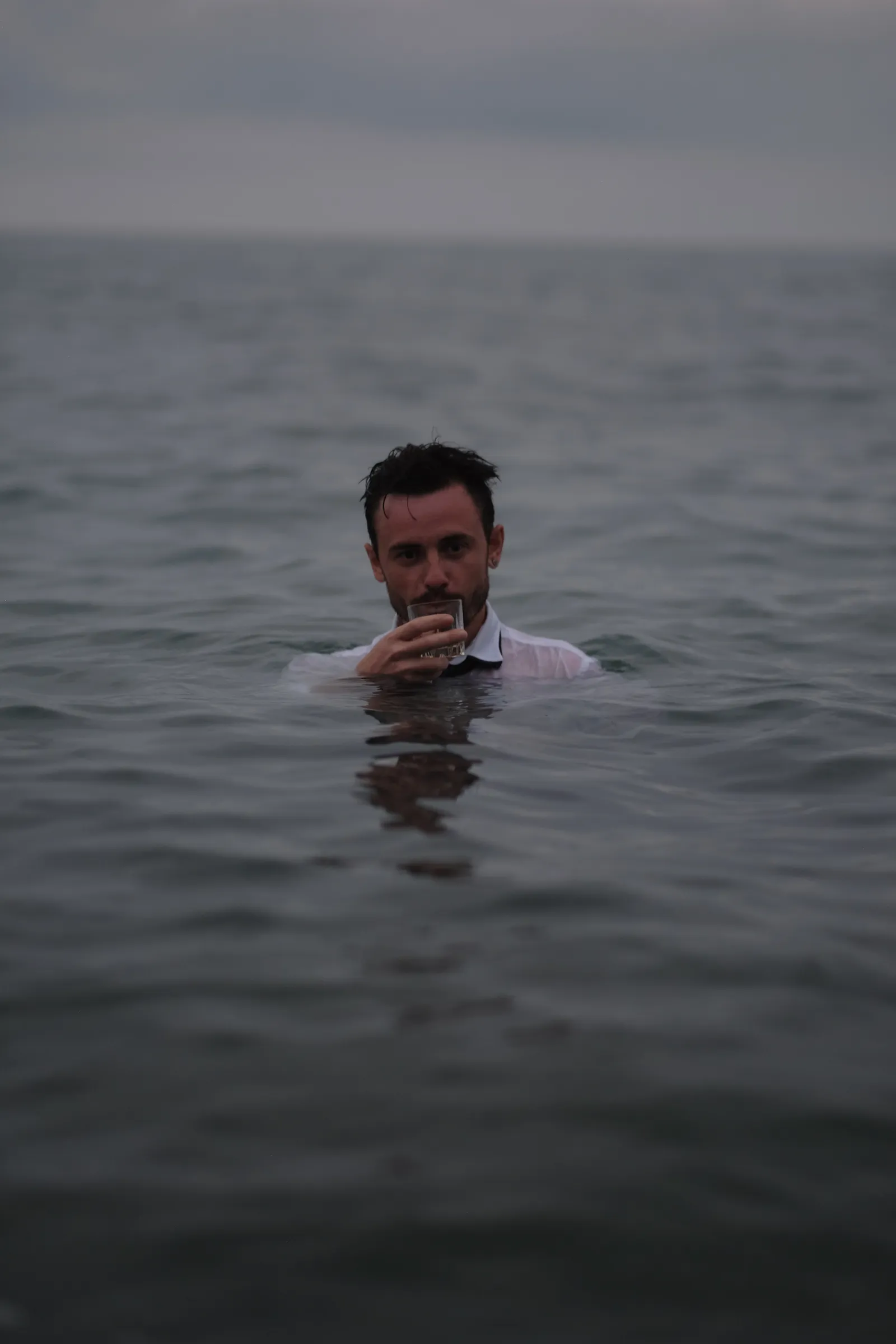 Man neck-deep in grey water, sipping from a glass