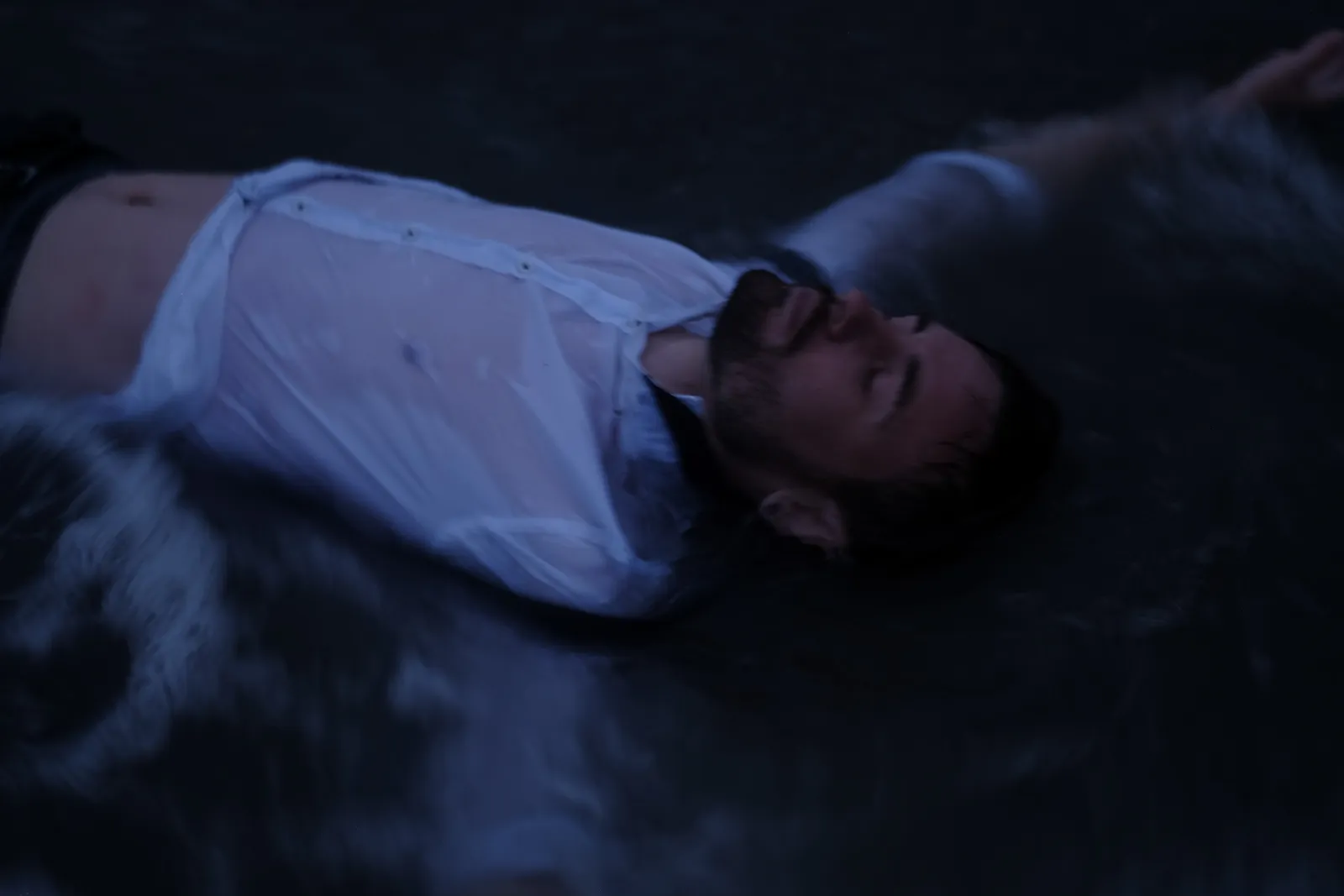 Man in a soaked white shirt lying on his back in the dark water, eyes closed