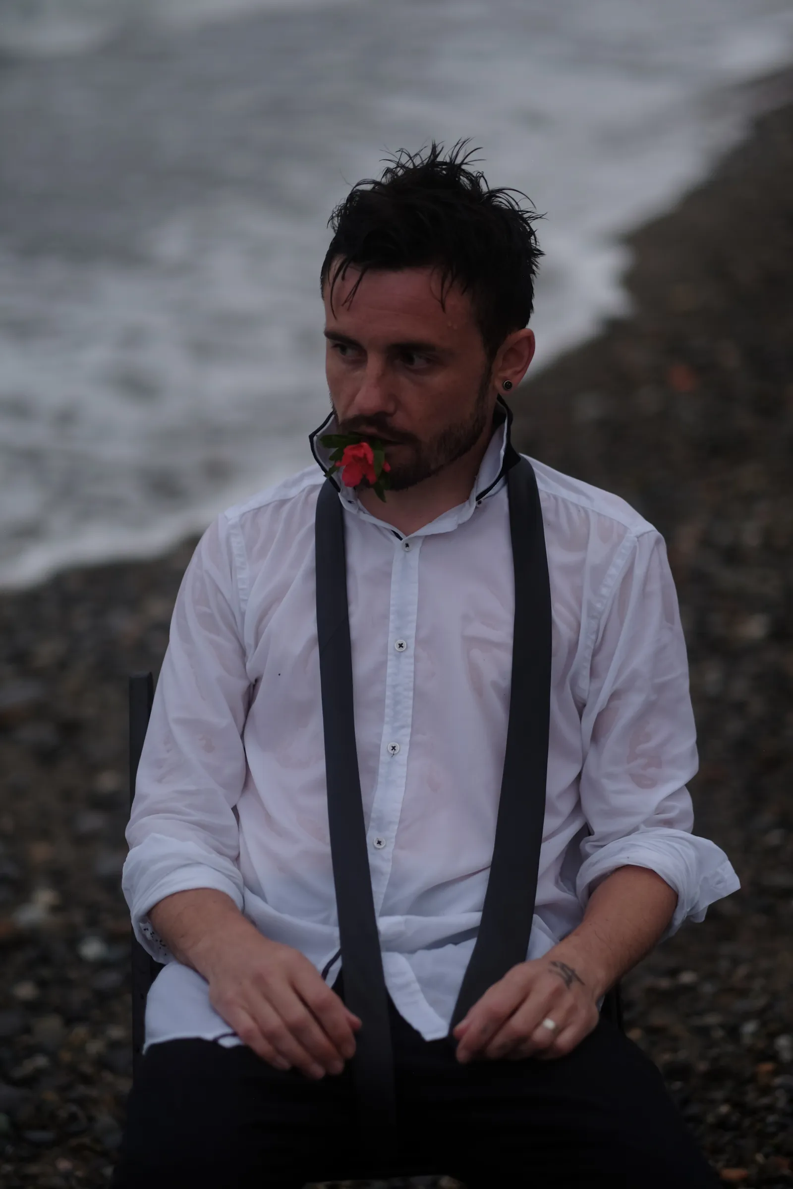 Soaked man in a white shirt and undone tie holding a red rose in his teeth