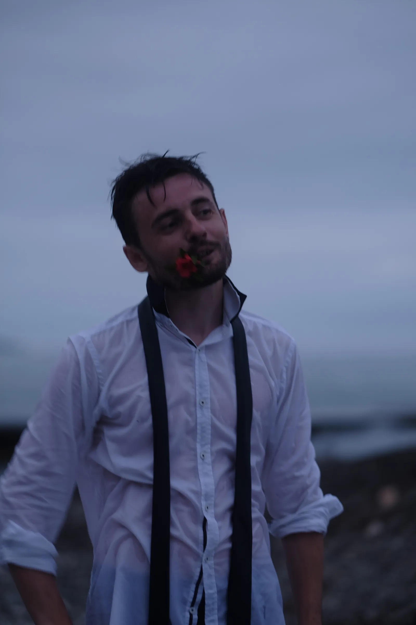 Drenched man with a rose in his teeth and an undone tie hanging loose, dusk sea behind