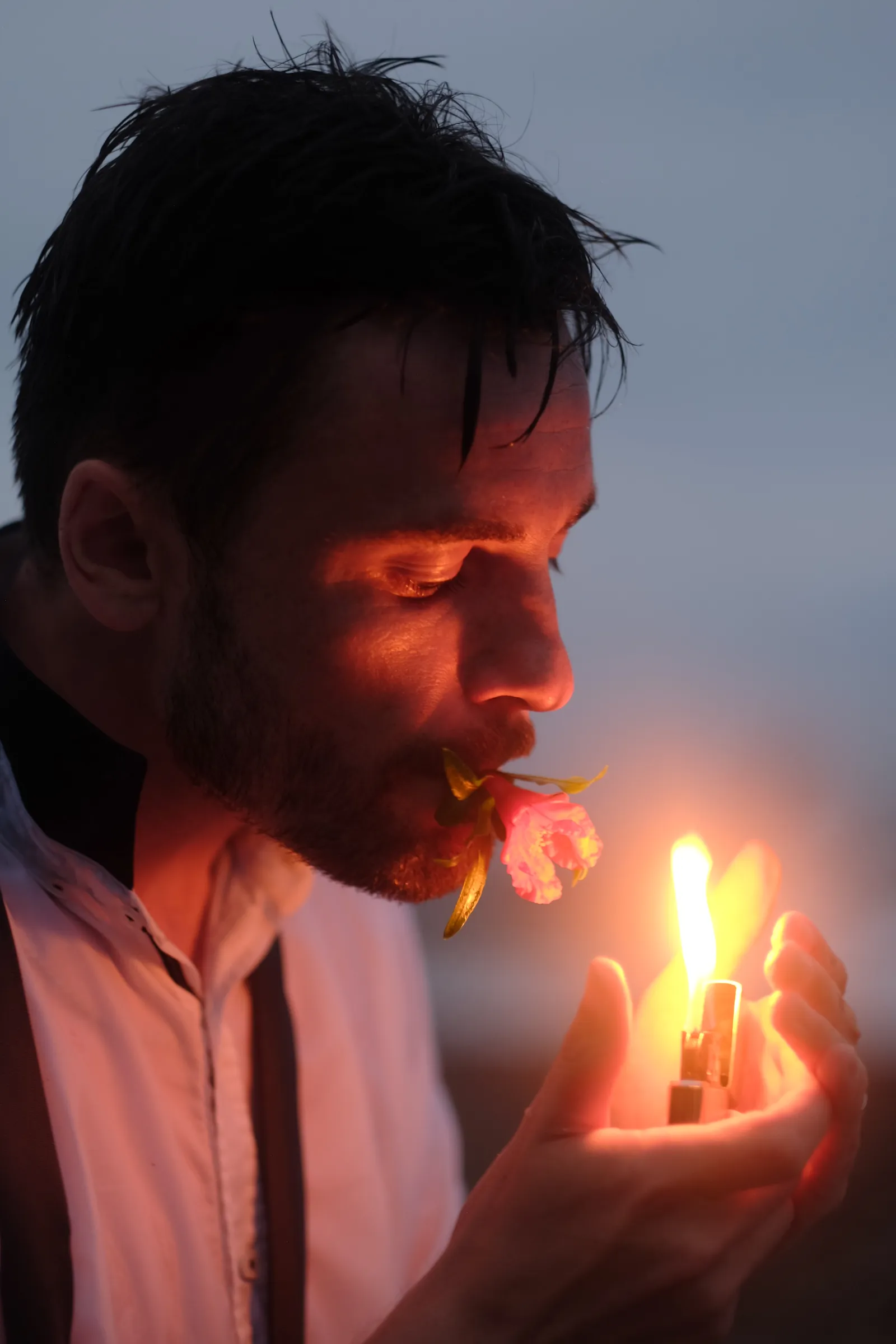 Man lighting a match beside a flower held in his mouth, flame lighting his face