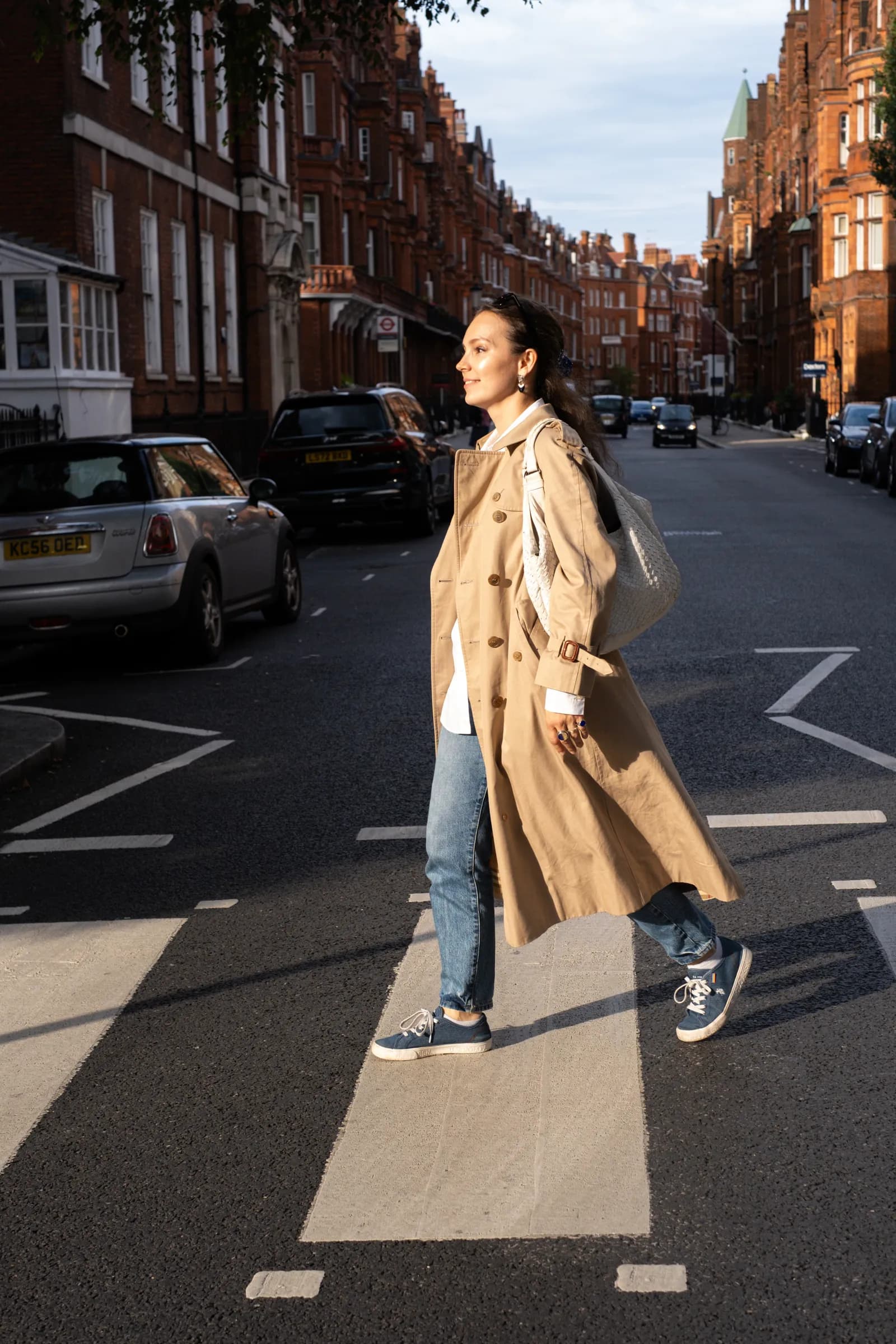 Woman in a trench coat crossing a sunlit red-brick street, bag over her shoulder