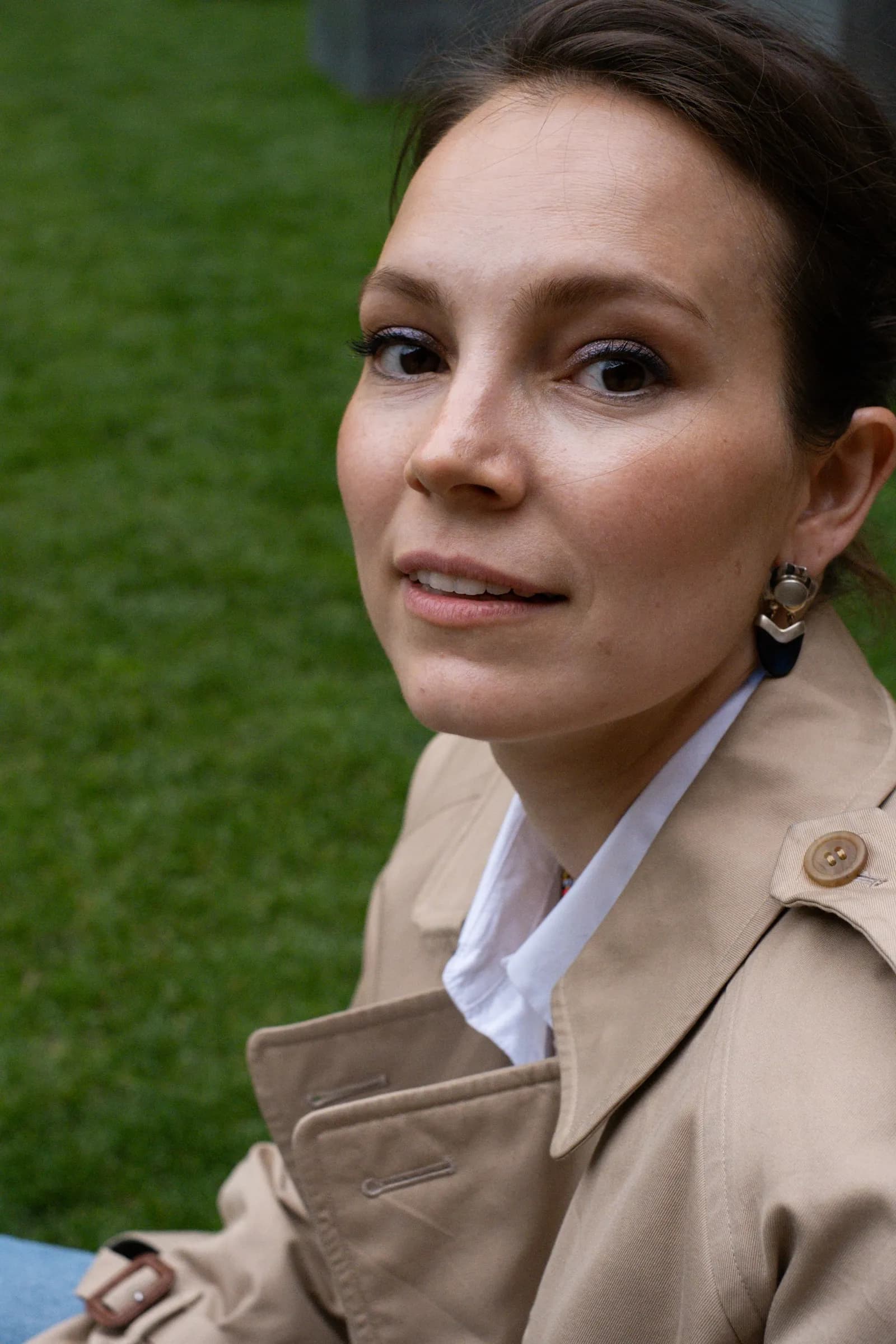 Close portrait of a woman in a trench coat over a white shirt, glancing back over her shoulder