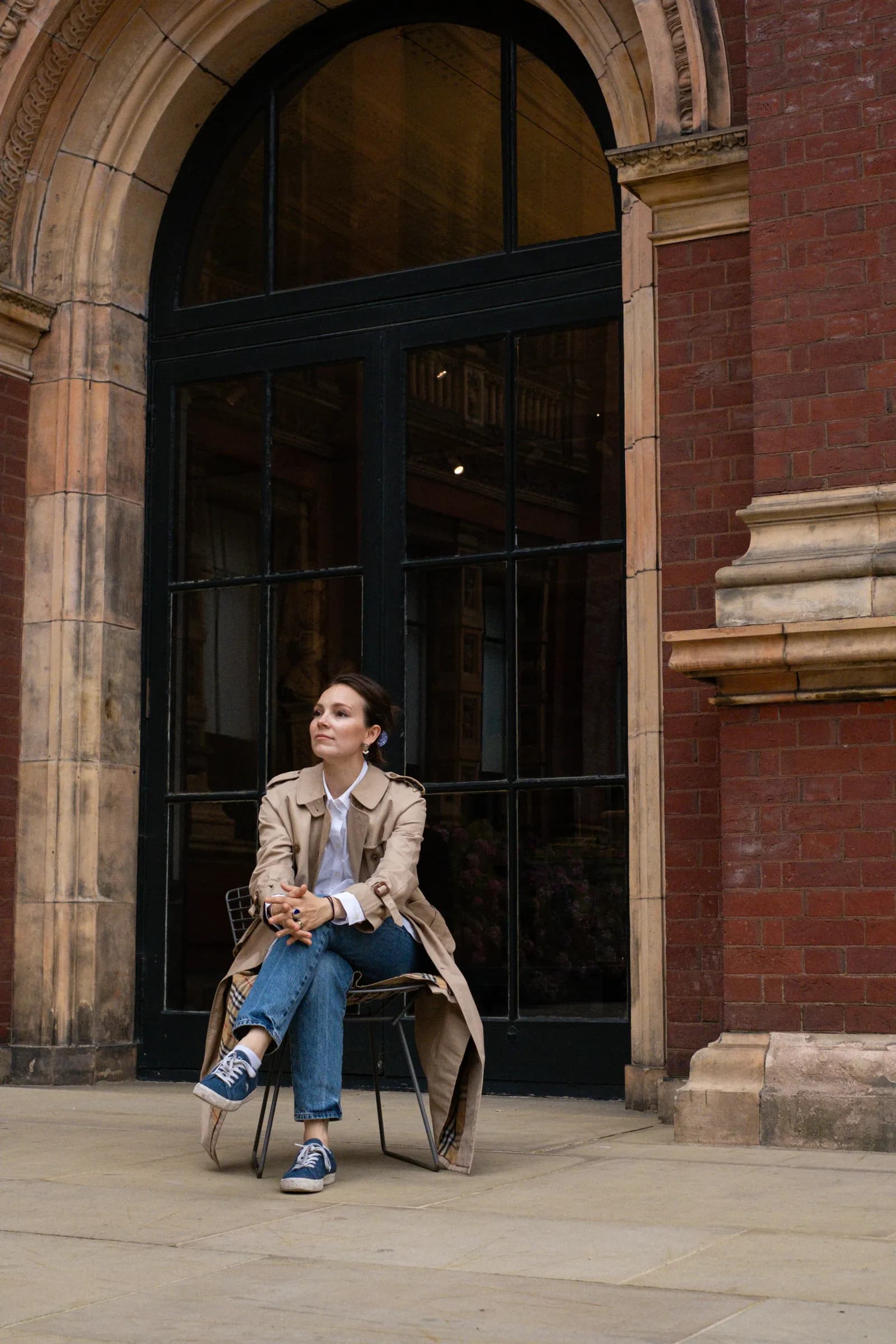 Woman in a trench coat sitting on a chair before the tall glass doors of a Victorian museum
