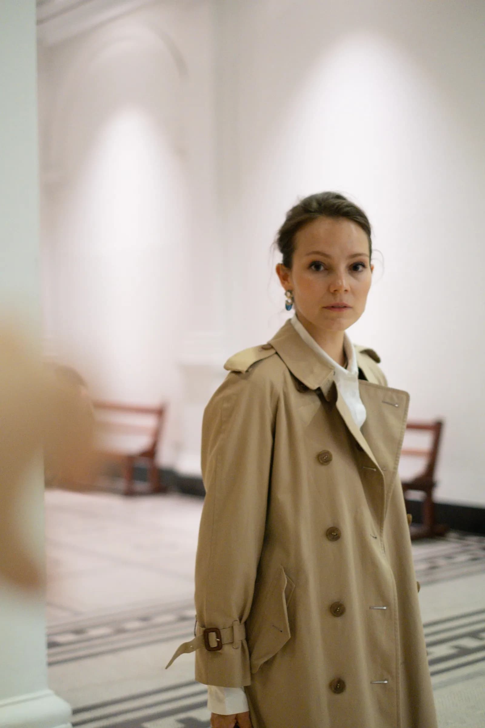 Woman in a trench coat standing in a white sculpture hall, caught mid-turn
