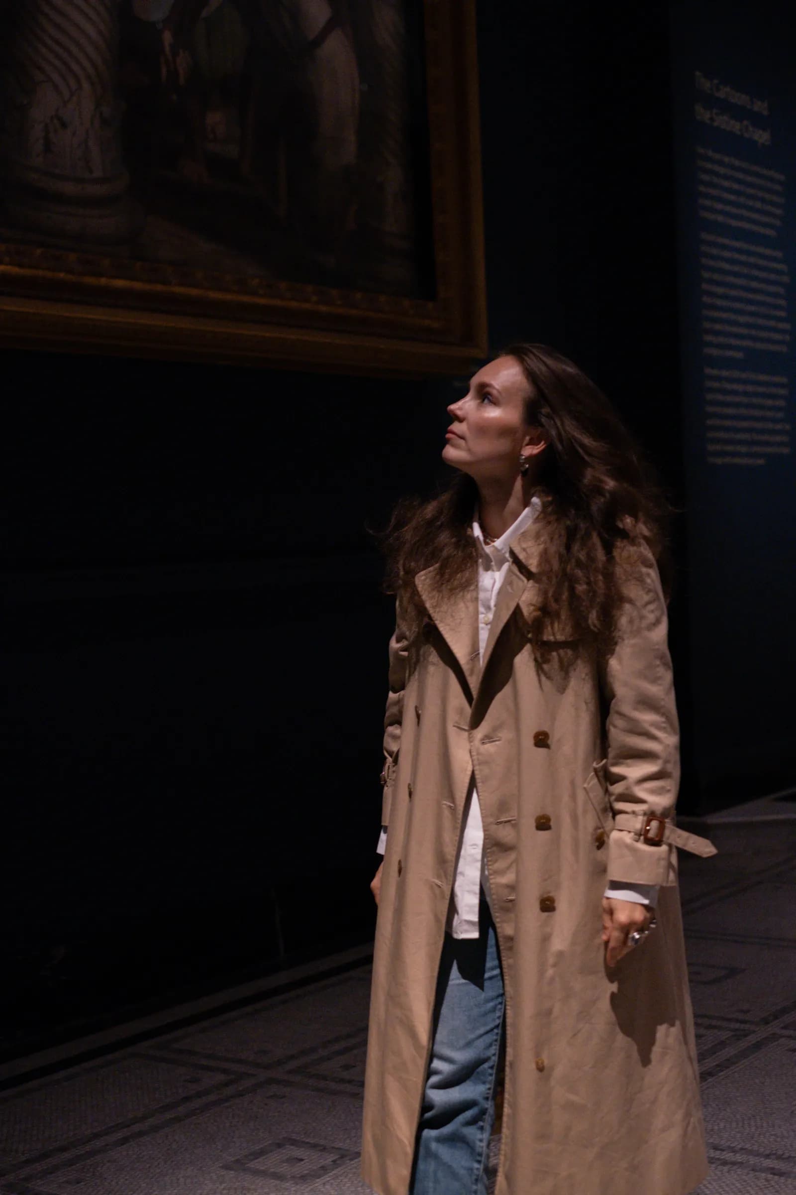 Woman in a trench looking up at a painting in a dim gallery