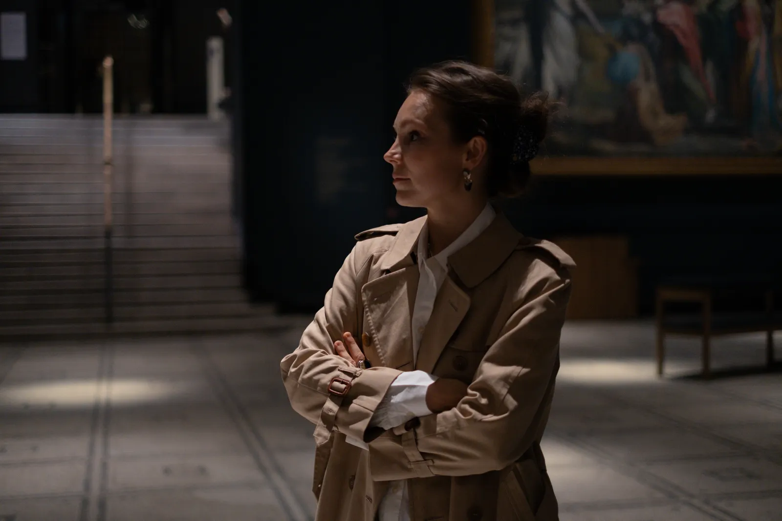 Woman with crossed arms in profile beneath the dark paintings of a museum wall