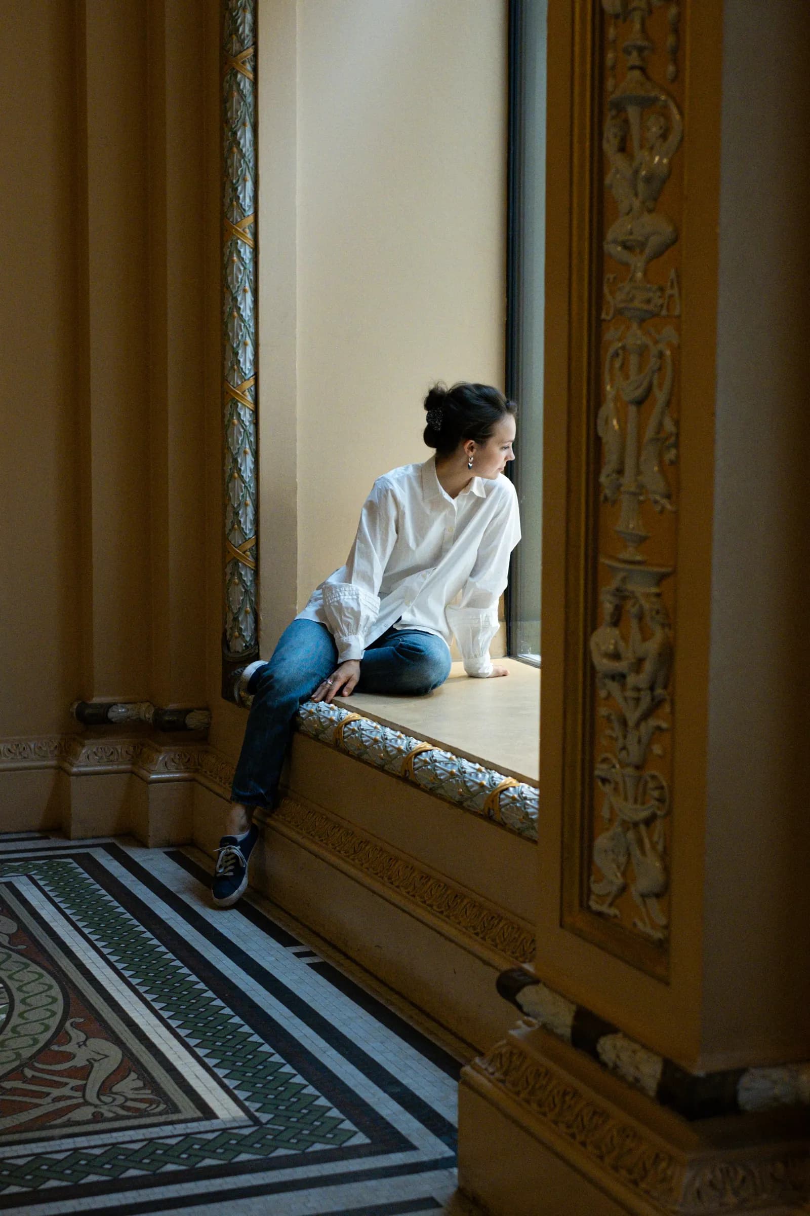 Woman sitting in a gilded window bay above a mosaic floor, looking out