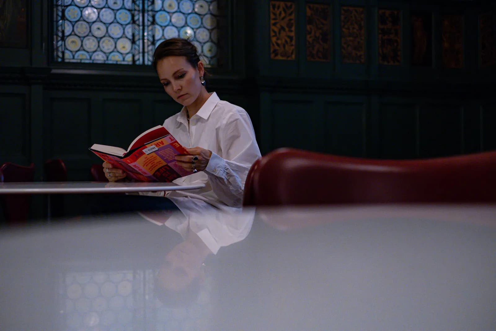 Woman reading a red-covered book at a table beneath stained-glass windows