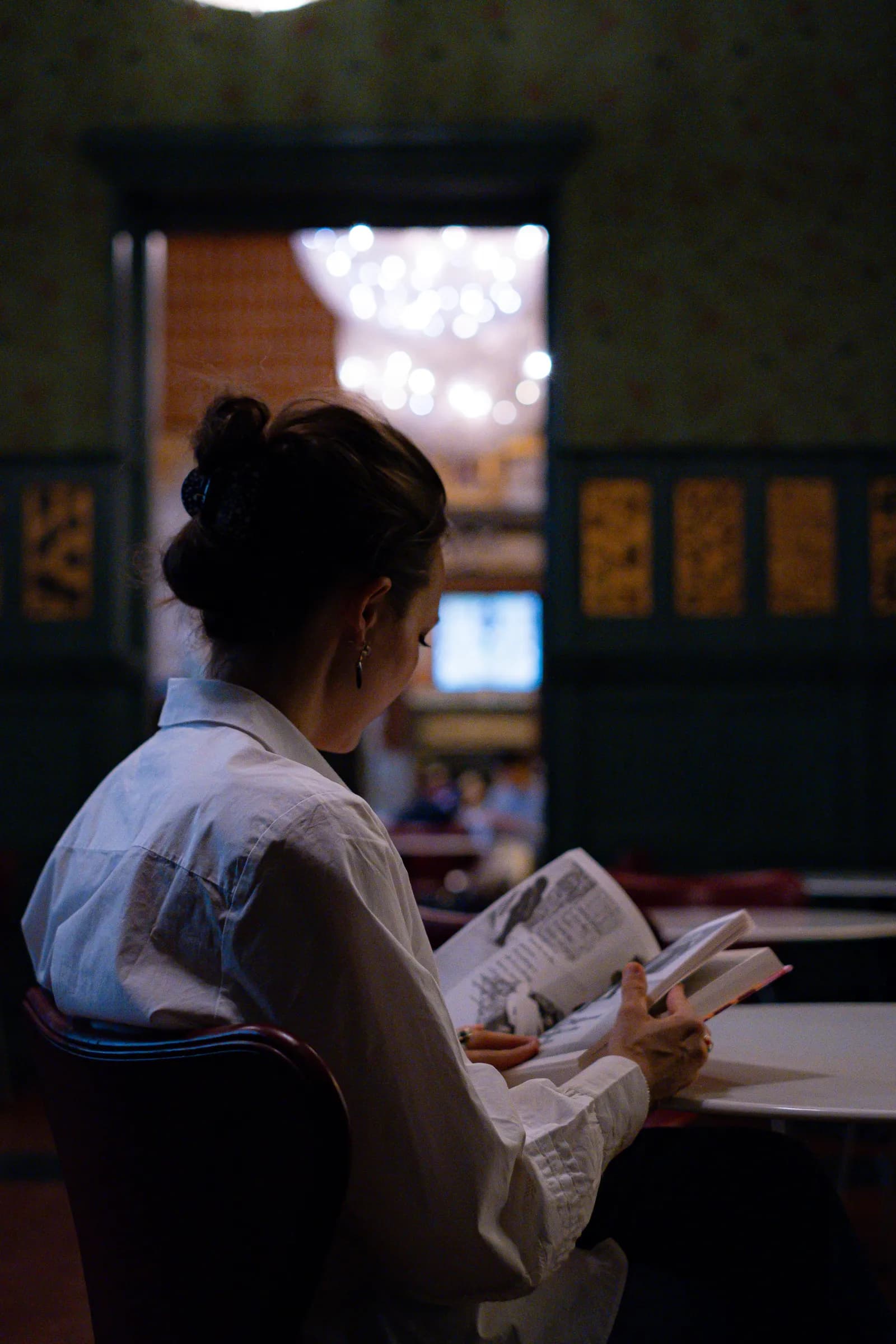 Woman seen from behind reading a newspaper in a dim panelled cafe, chandelier glowing beyond