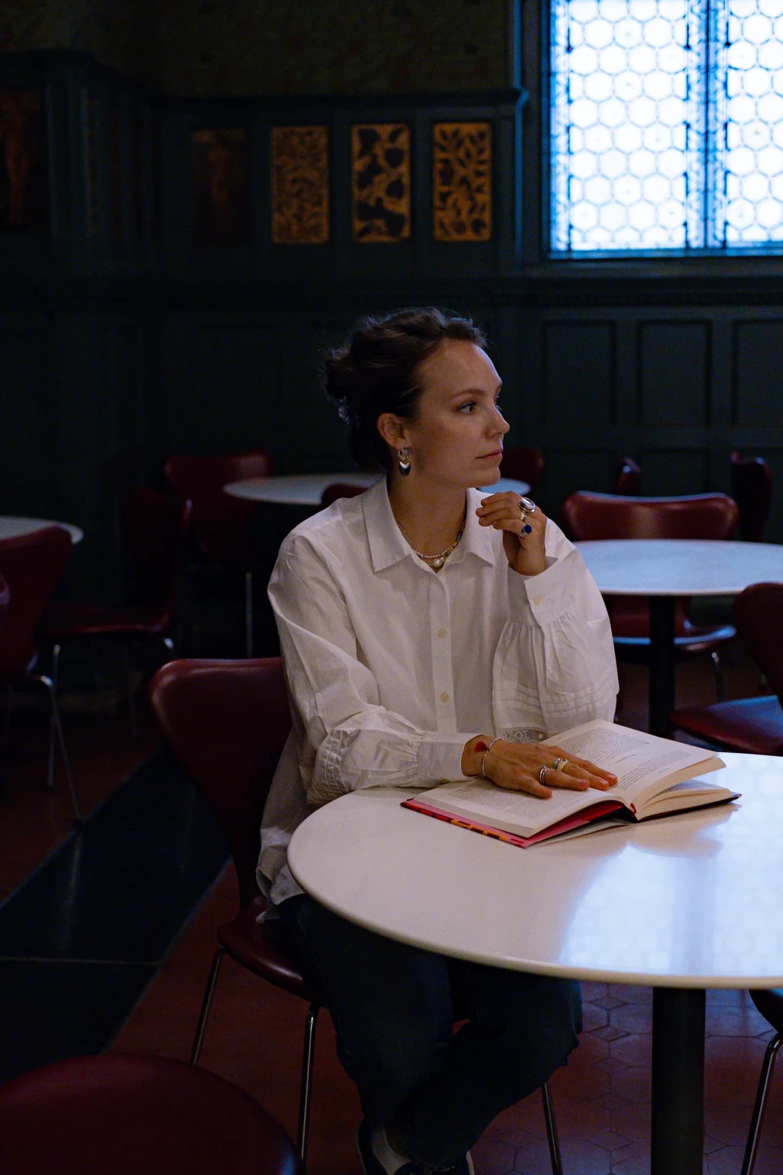 Woman at a museum cafe table with an open book, chin on her hand, looking to the window