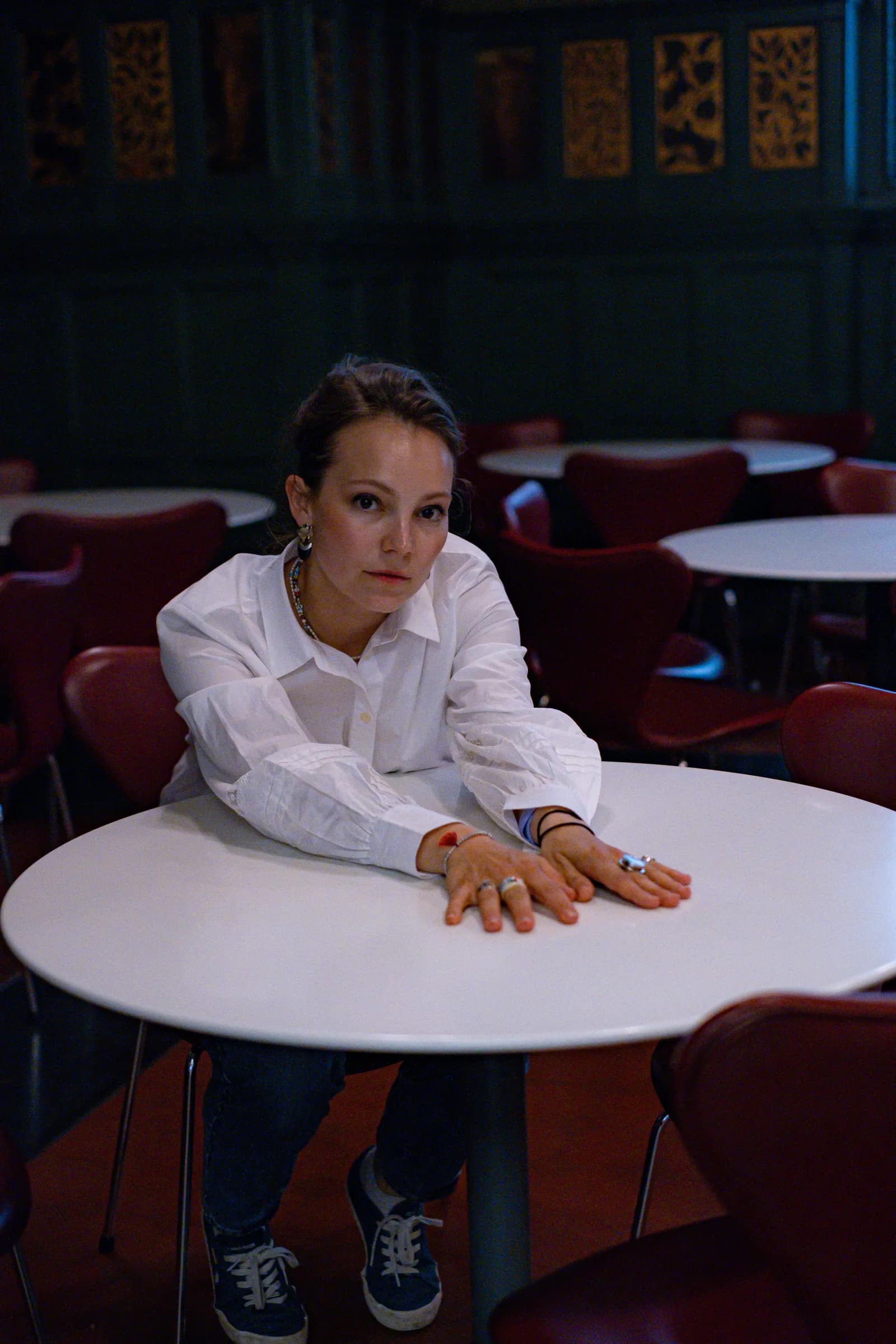 Woman in a white shirt leaning across a cafe table, ringed hands folded, looking into the lens