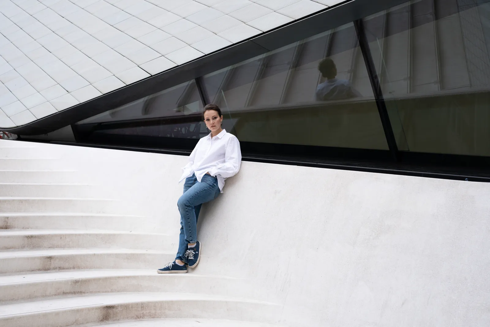 Woman in a white shirt and jeans reclining on sweeping white concrete steps under a modern glass roof