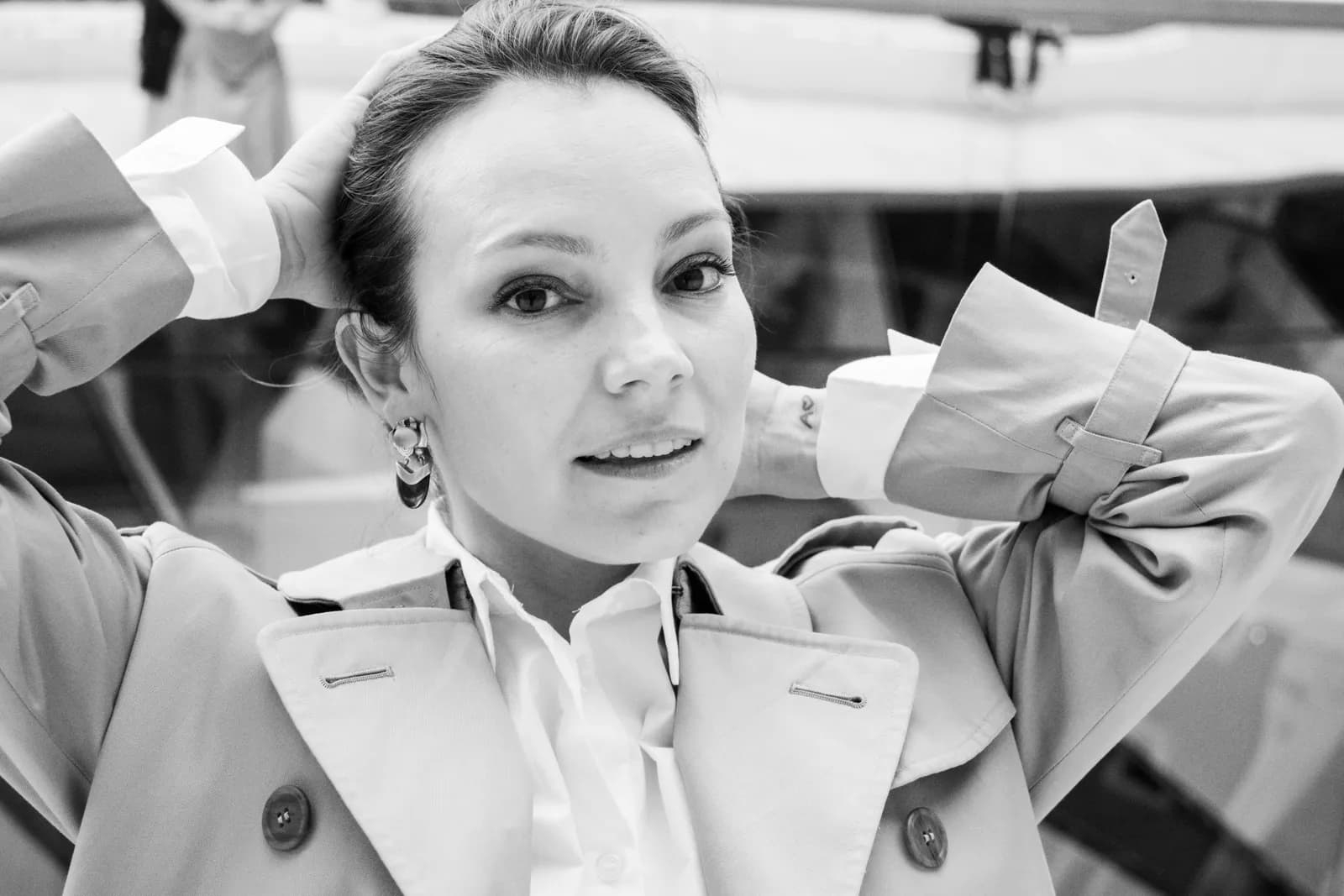 Black and white portrait, hands raised behind her head, trench collar open
