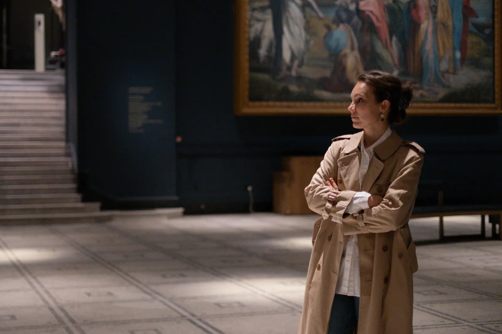 Woman in a trench coat with crossed arms in a museum hall, a vast painting behind her