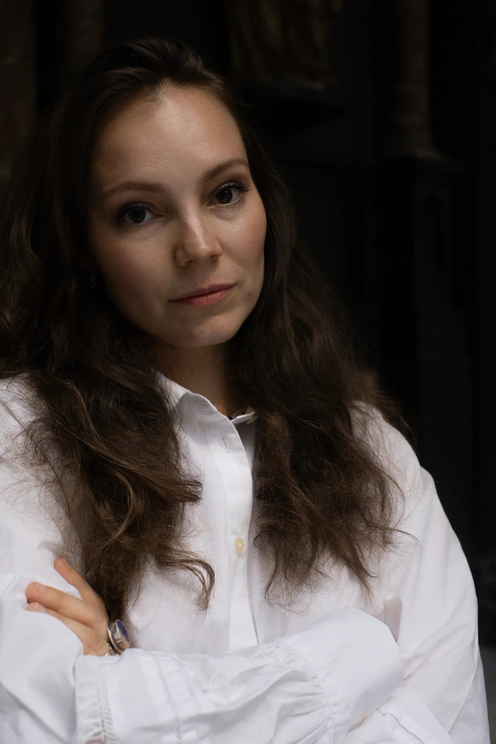 Close portrait with crossed arms in a white shirt, dark drapery behind