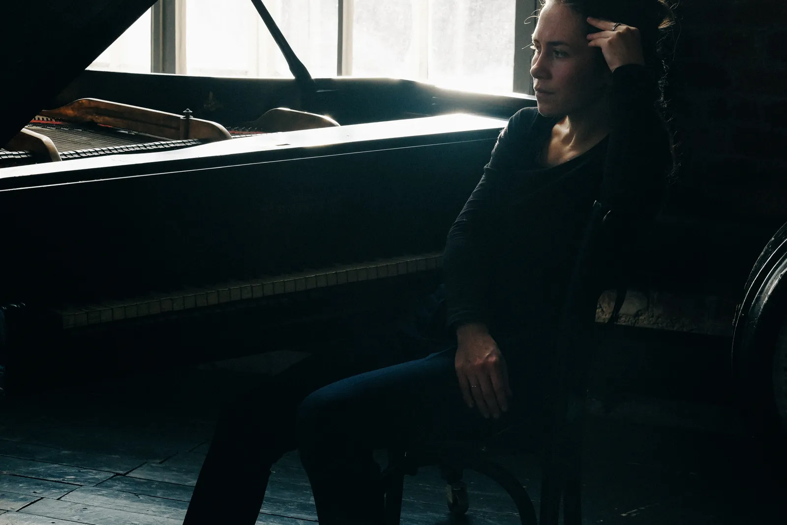 Woman seated by a grand piano at a tall window, hand raised to her head