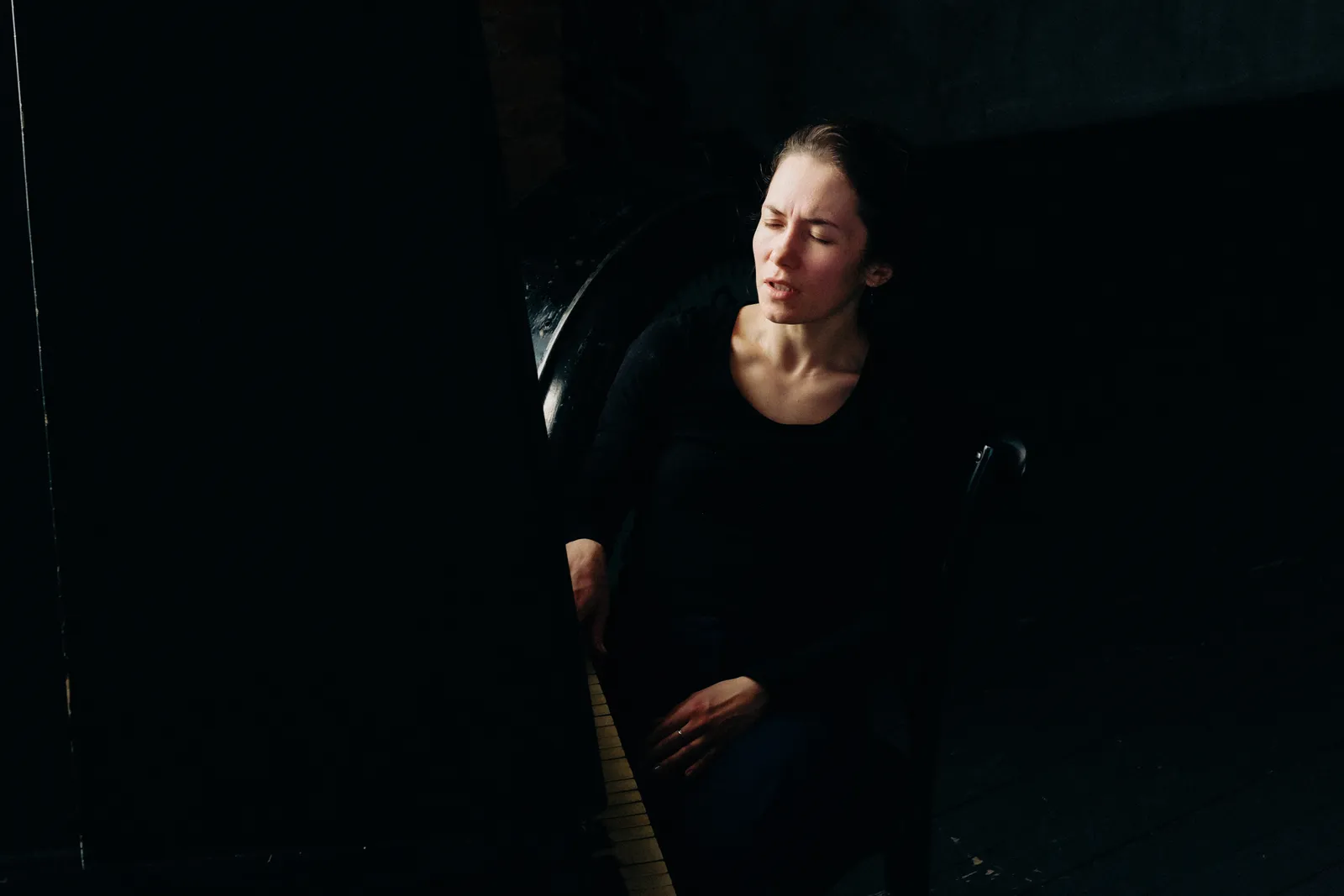 Woman in black seated by the open piano lid, face lifted into the light, eyes closed