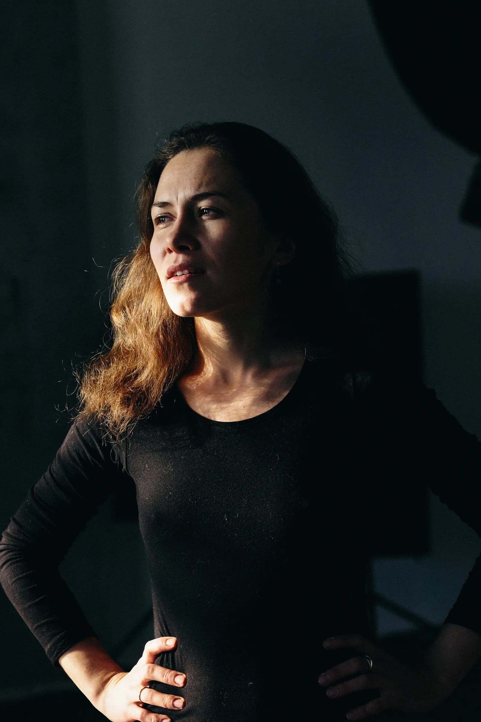 Woman in black standing in a beam of hard light, hands on hips, looking off into the dark