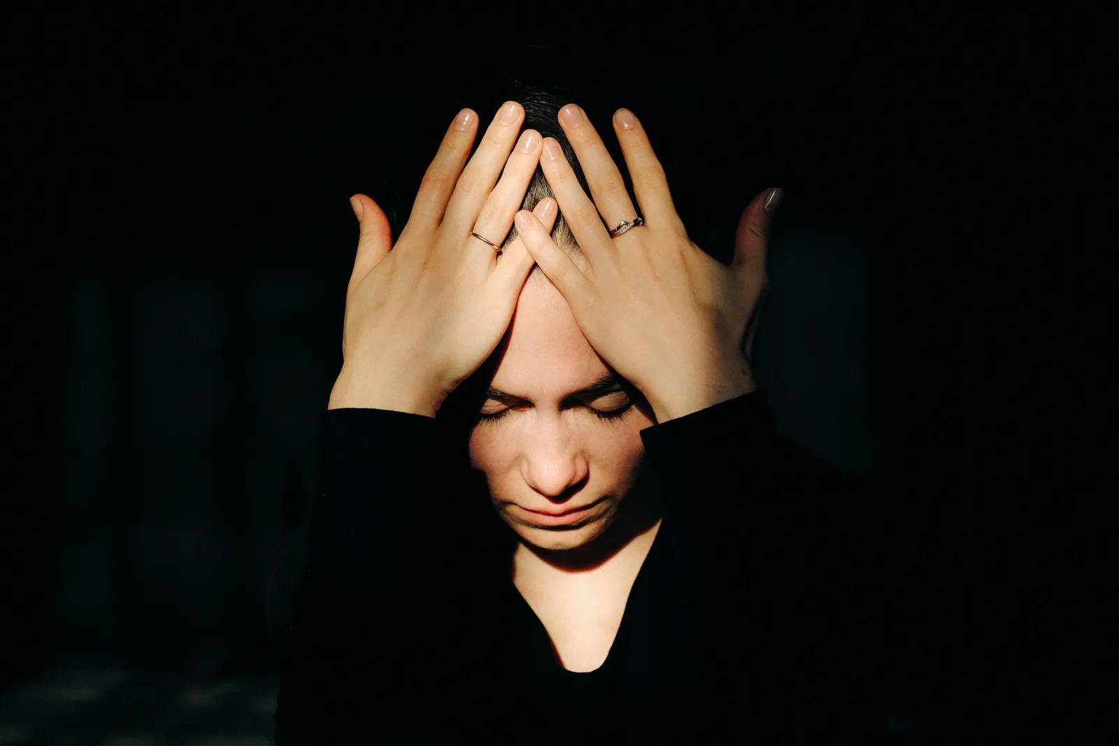 Woman with both hands pressed to her forehead, ringed fingers lit against the dark